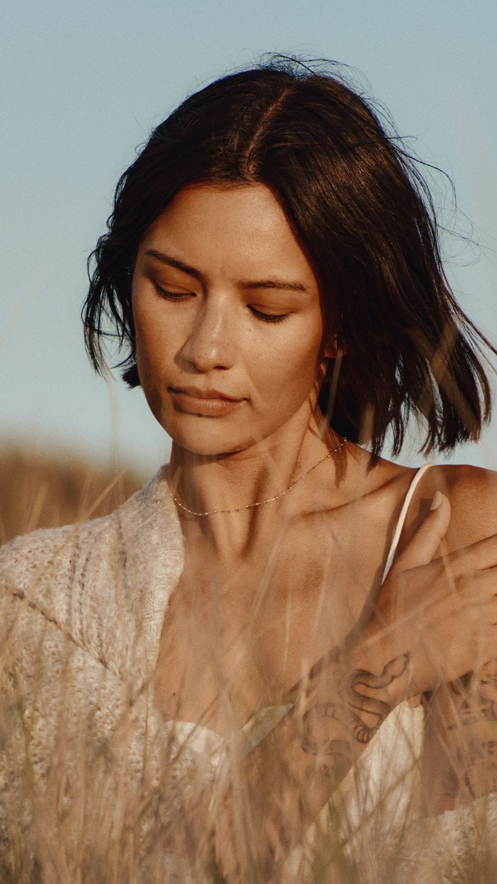 A woman with dark hair, wearing a white top and light cardigan, looks down with her tattooed arm crossed over her chest, surrounded by tall dry grass against a blue sky.