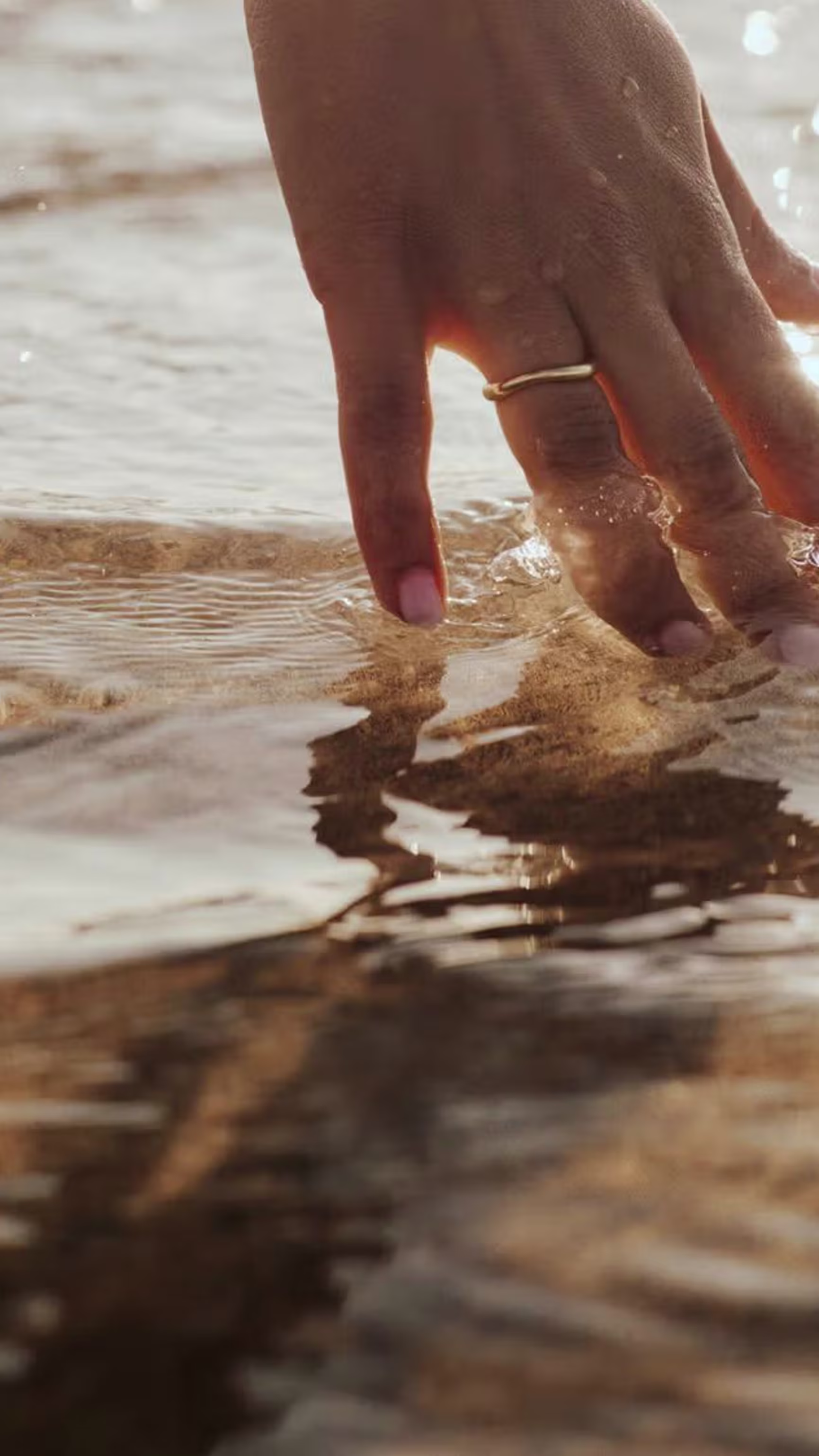 A hand with a gold ring gently touches sparkling water.