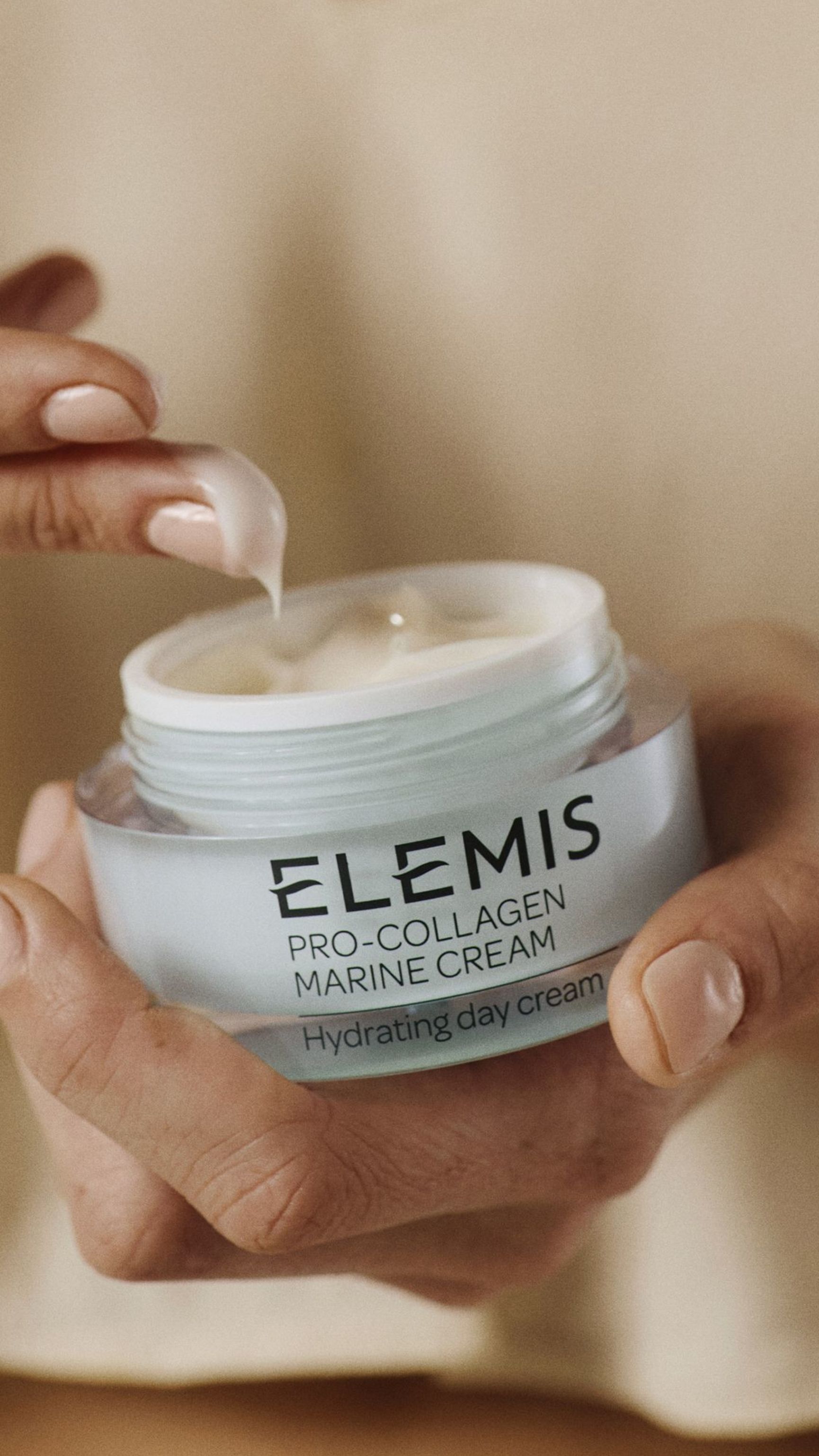 Hands holding an open jar of Elemis Pro-Collagen Marine Cream, scooping out product.