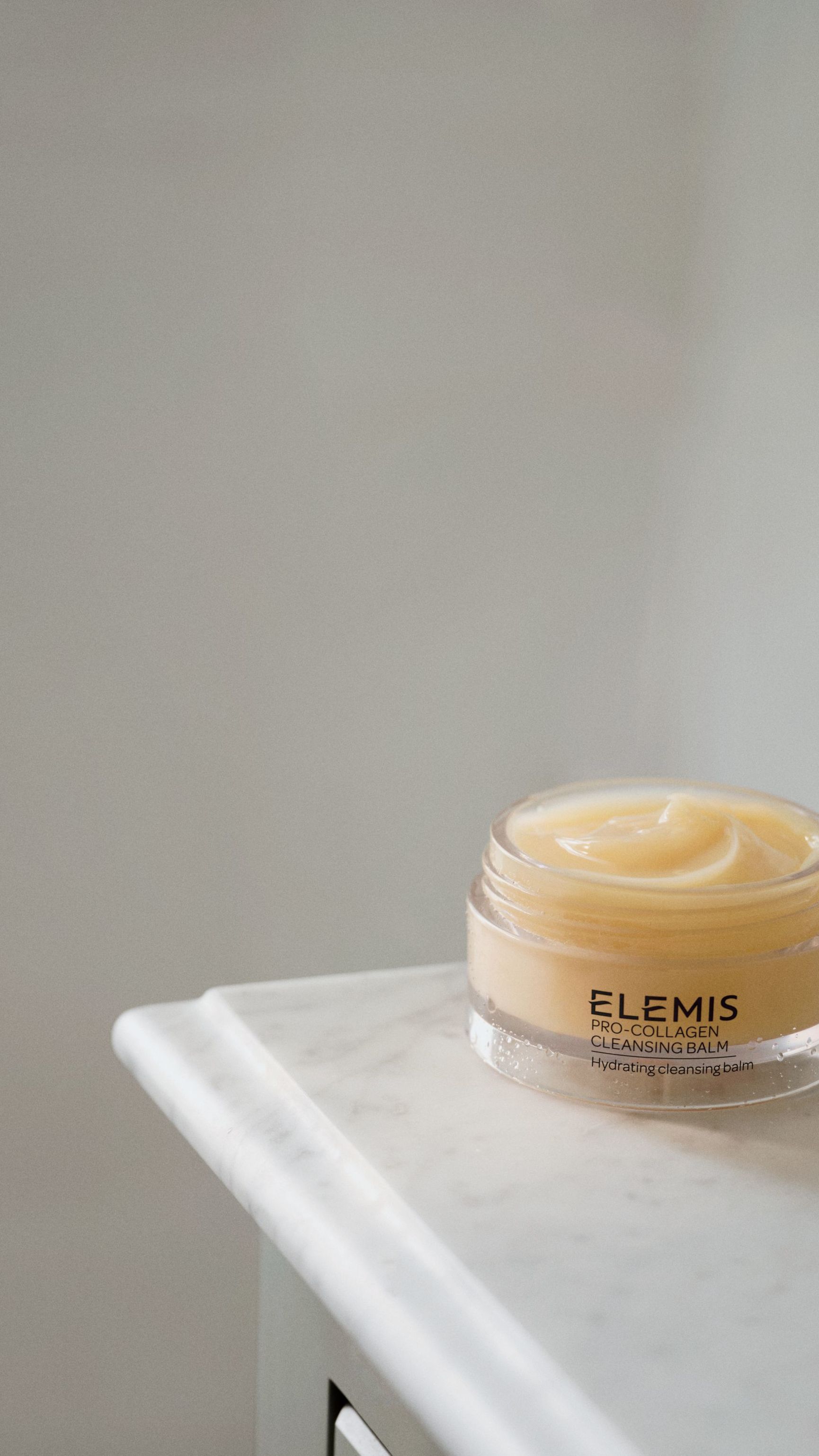 A woman lying in tall grass, holding a jar of Elemis Pro-Collagen Cleansing Balm under a blue sky.