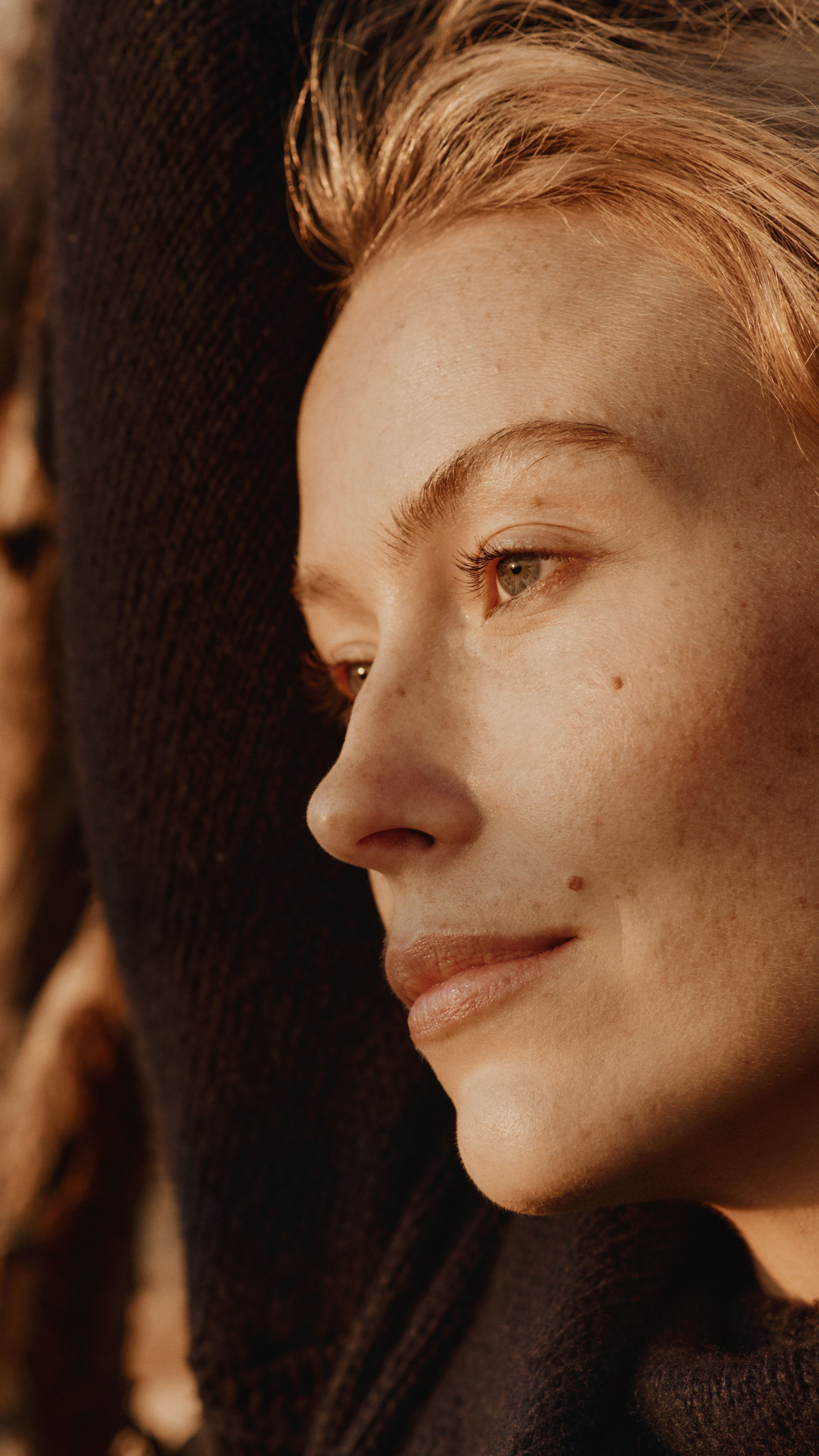 Close-up profile of a woman with blonde hair and freckles, wearing a dark sweater, illuminated by warm light with wooden logs in the background.