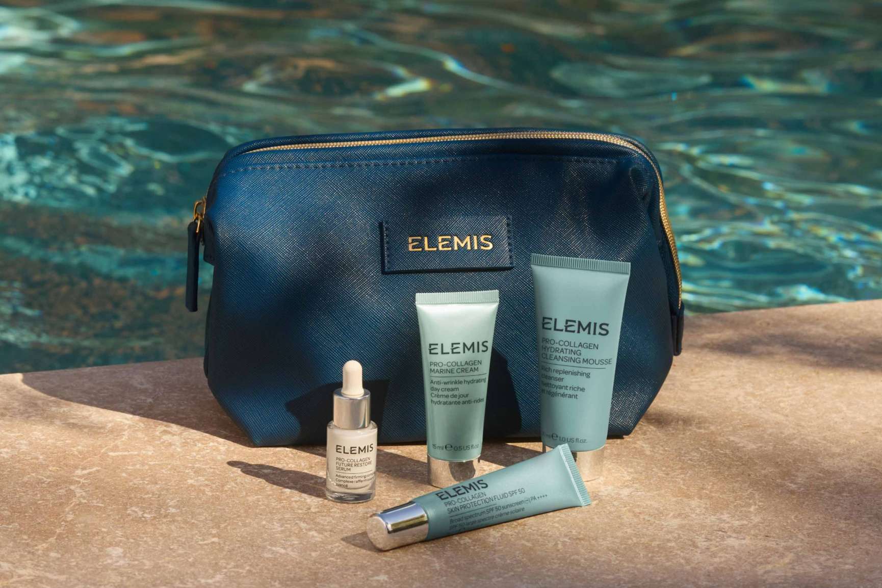 A navy Elemis cosmetic bag and Pro-Collagen skincare products sit on a stone surface next to a swimming pool.