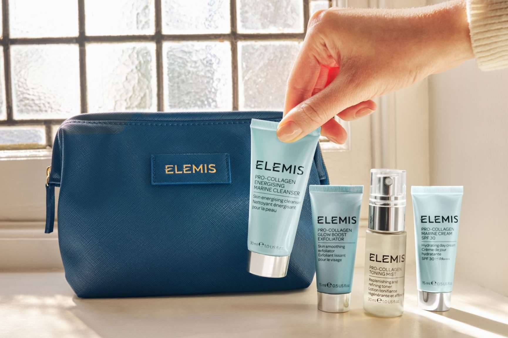 A hand holds an Elemis cleanser next to three other Elemis skincare products and a dark blue Elemis cosmetics bag.