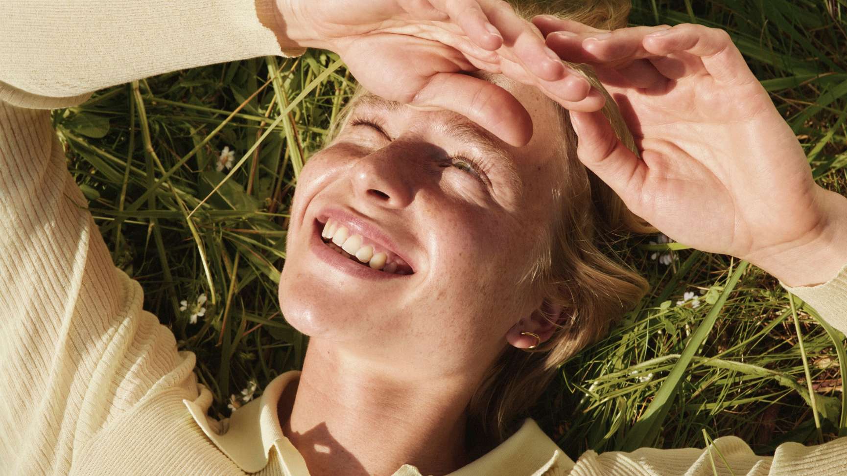 A person with freckles lies in green grass, smiling and shading their eyes from the sun.