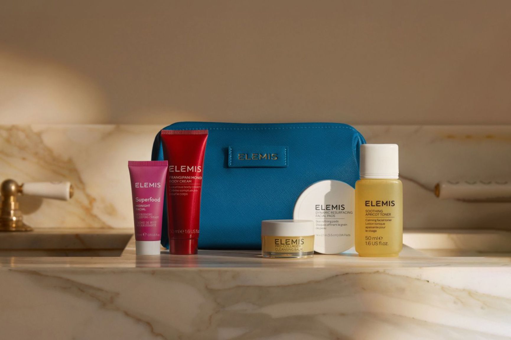 A blue Elemis cosmetic bag sits behind five Elemis skincare products: a pink tube, a red tube, a small white jar, a round white container, and a yellow bottle.