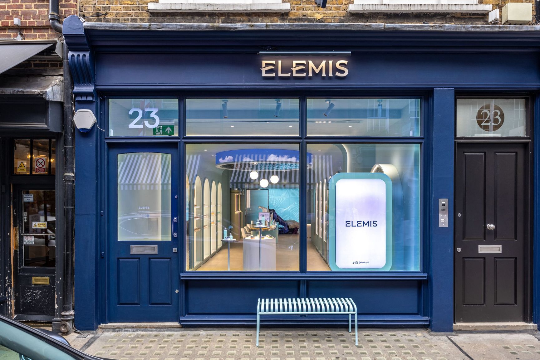 Exterior de una tienda Elemis con fachada azul oscuro, un gran escaparate y un banco azul claro.