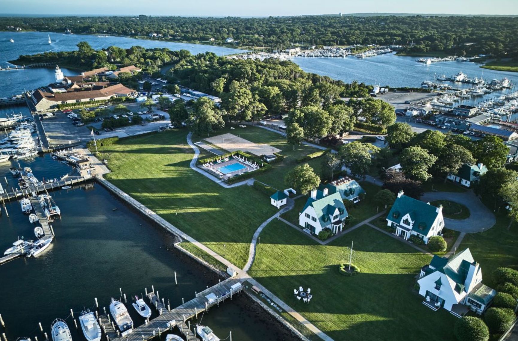 Aerial view of a sprawling waterfront resort with white buildings, green roofs, a pool, volleyball court, extensive lawns, and multiple boat-filled marinas.