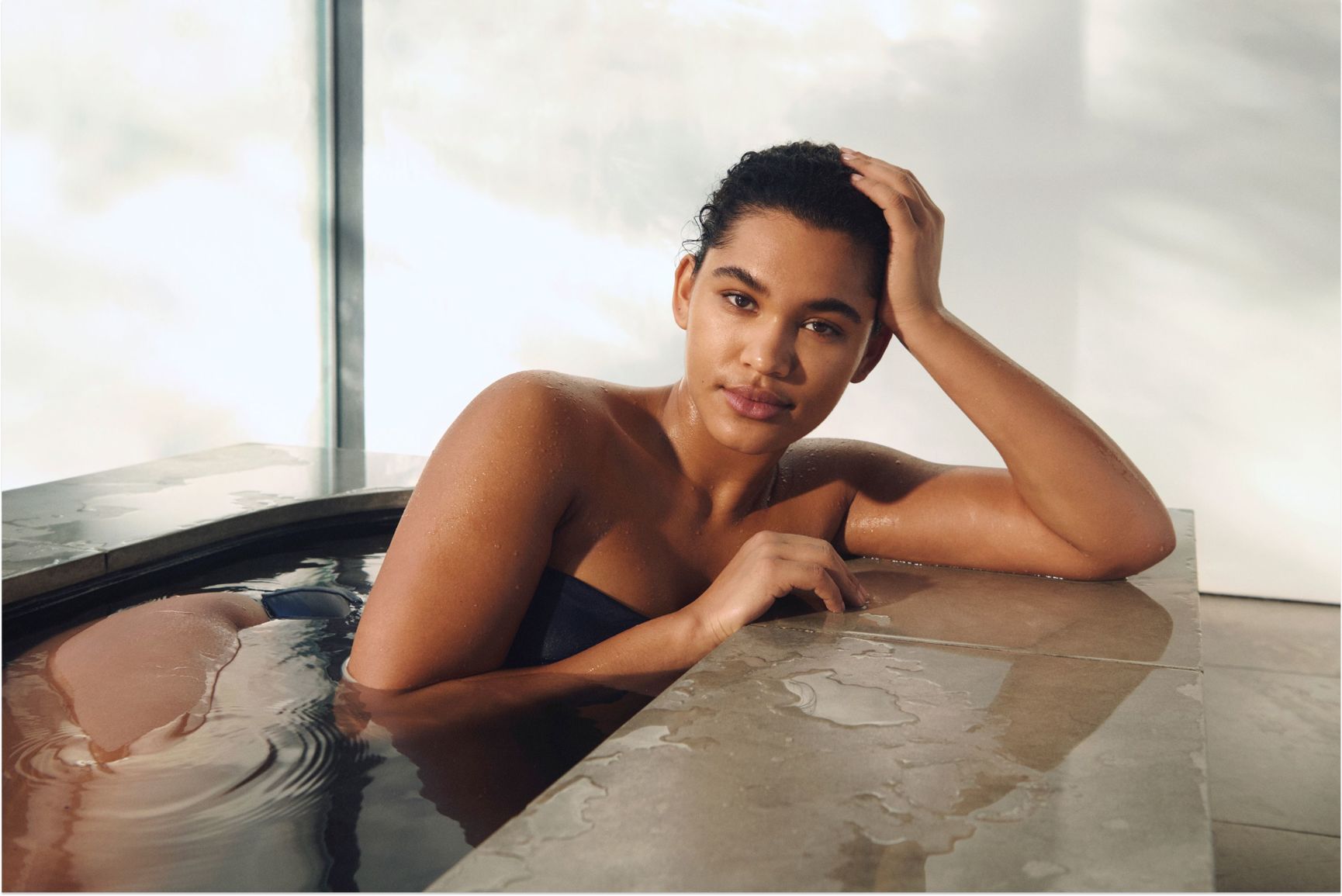 A woman with wet hair and skin in a bath, leaning on the tiled edge and looking at the camera.