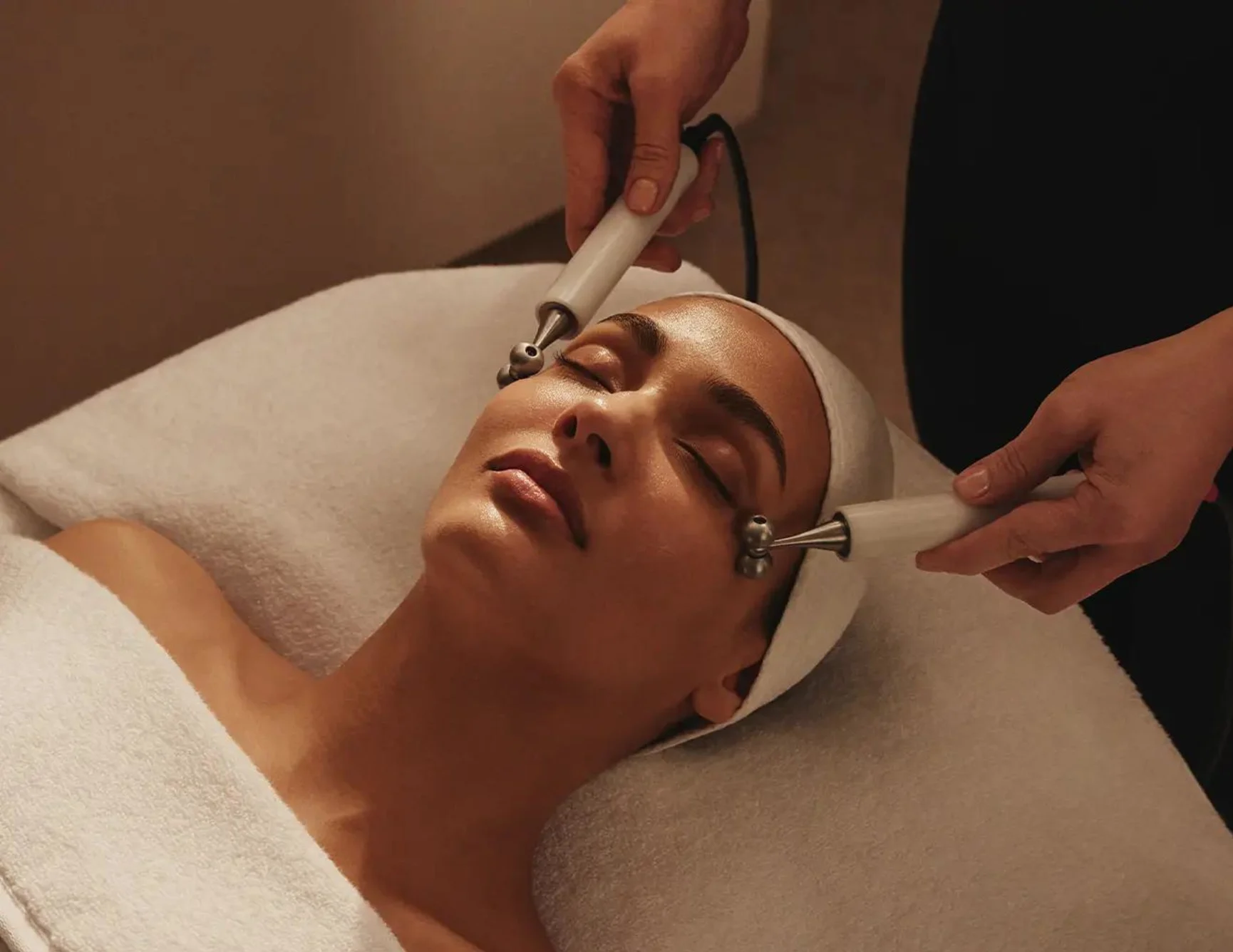 A woman receives a facial massage, with several skincare capsules on the white bedsheet.