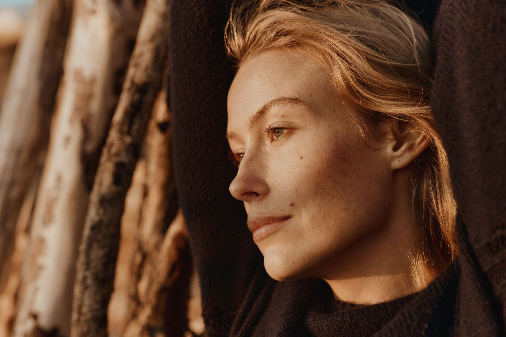 Close-up profile of a woman with blonde hair and freckles, wearing a dark sweater, illuminated by warm light with wooden logs in the background.