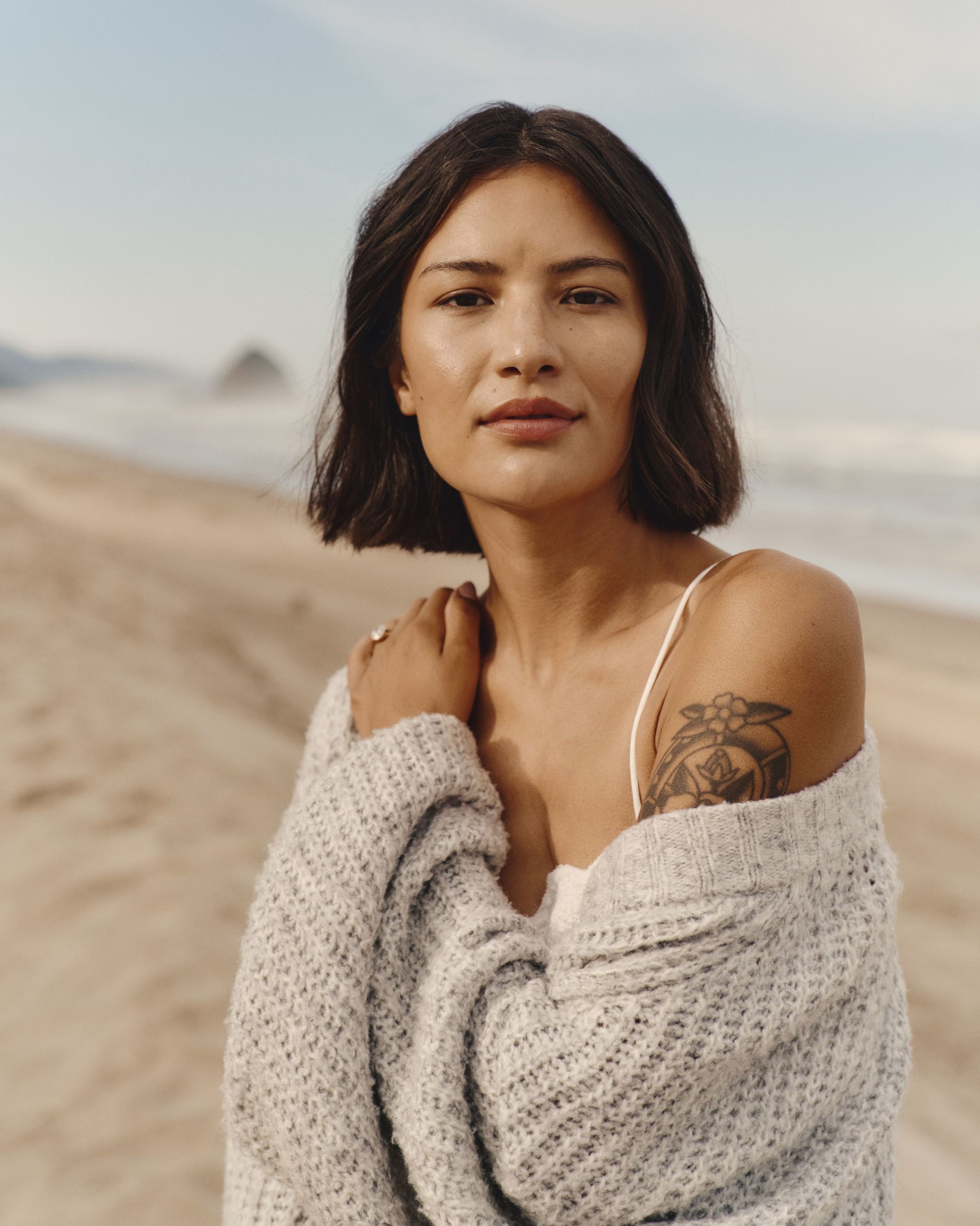 A woman with short dark hair and a tattoo wears a grey cardigan on a beach with a distant rock formation.