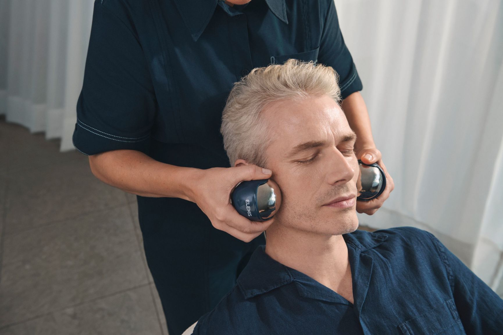 A woman receives a facial massage, with several skincare capsules on the white bedsheet.