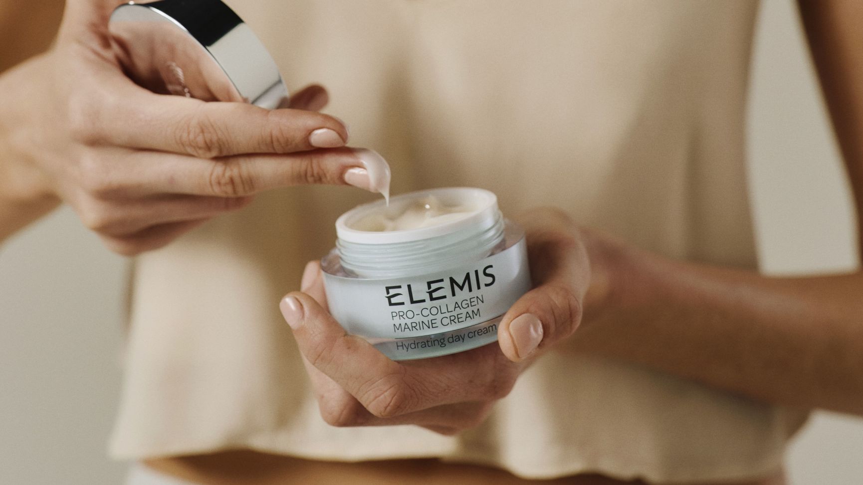 Hands holding an open jar of Elemis Pro-Collagen Marine Cream, scooping out product.