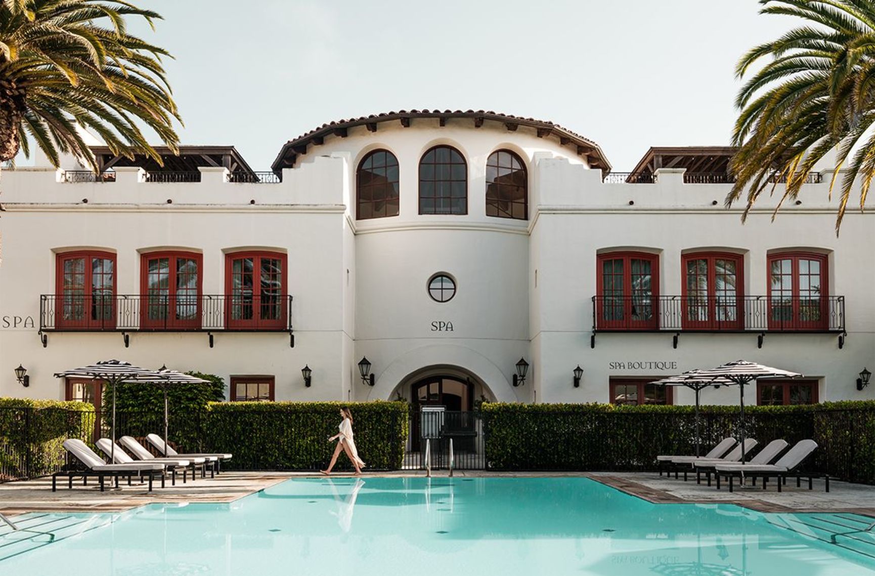 A white spa building with red accents overlooks a swimming pool where a person in a robe walks.