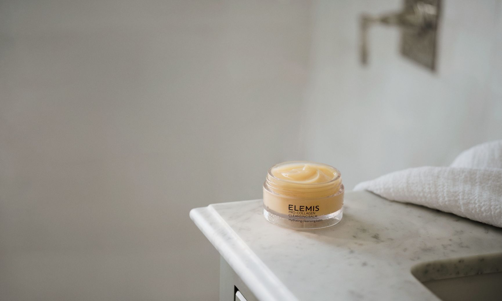 A woman lying in tall grass, holding a jar of Elemis Pro-Collagen Cleansing Balm under a blue sky.