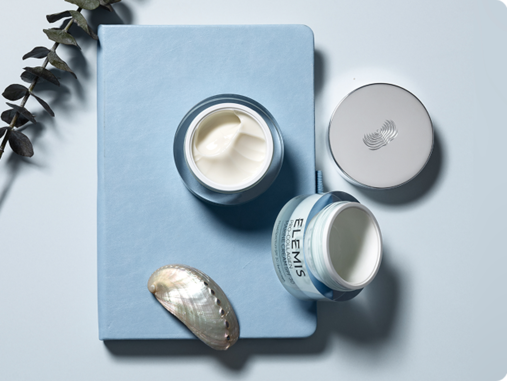 Open jar of Elemis Pro-Collagen Marine Cream next to an abalone shell and eucalyptus leaves on a blue notebook.