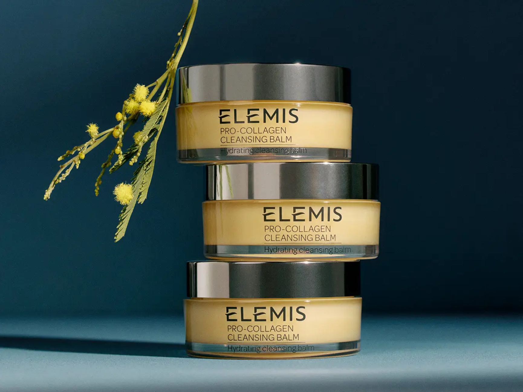 Three stacked jars of Elemis Pro-Collagen Cleansing Balm next to a sprig of yellow mimosa flowers.