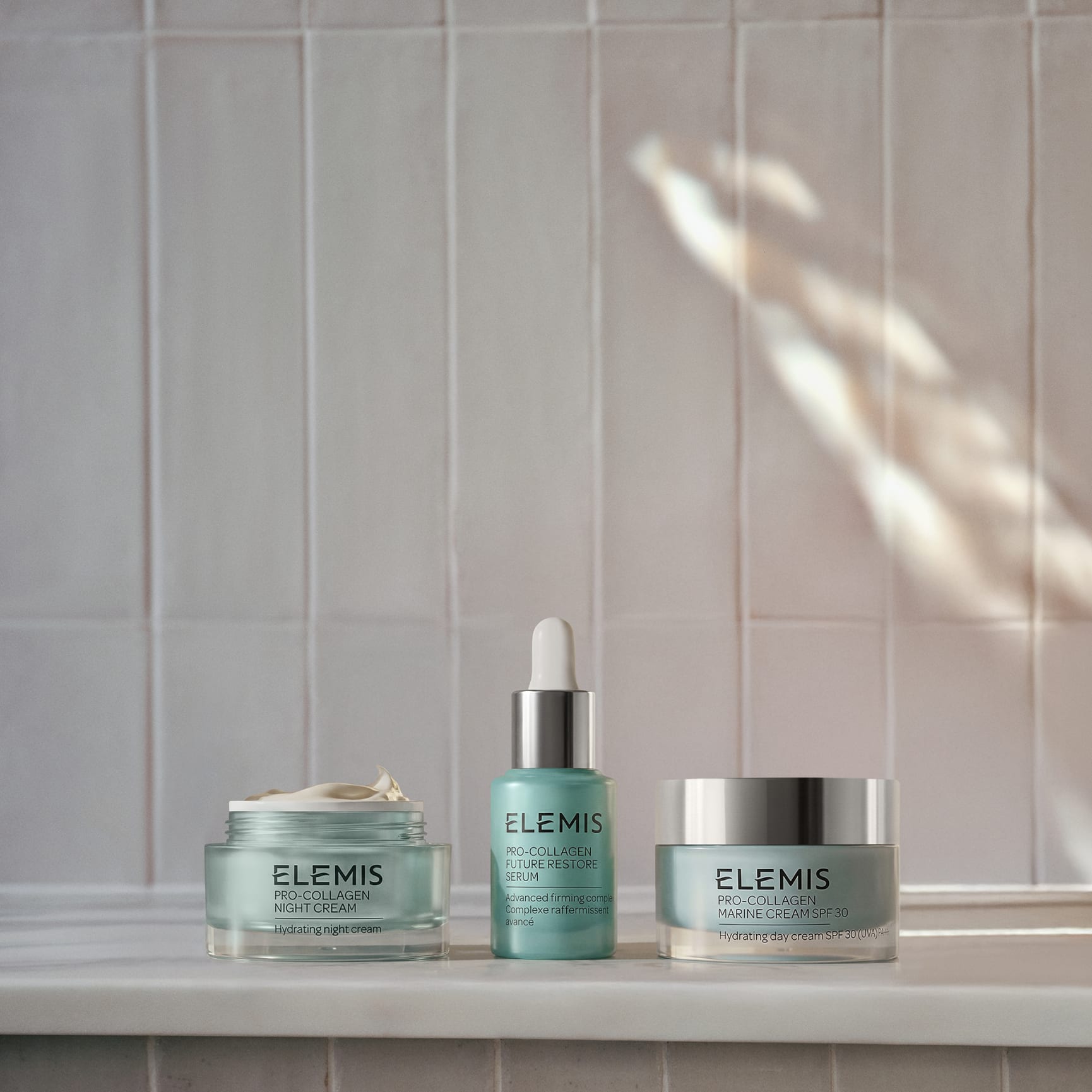Three ELEMIS Pro-Collagen products arranged on a bathroom ledge against pale tiled wall: an open Pro-Collagen Night Cream jar, Pro-Collagen Elixir serum with dropper, and closed Pro-Collagen Marine Cream SPF jar, lit by soft natural light.