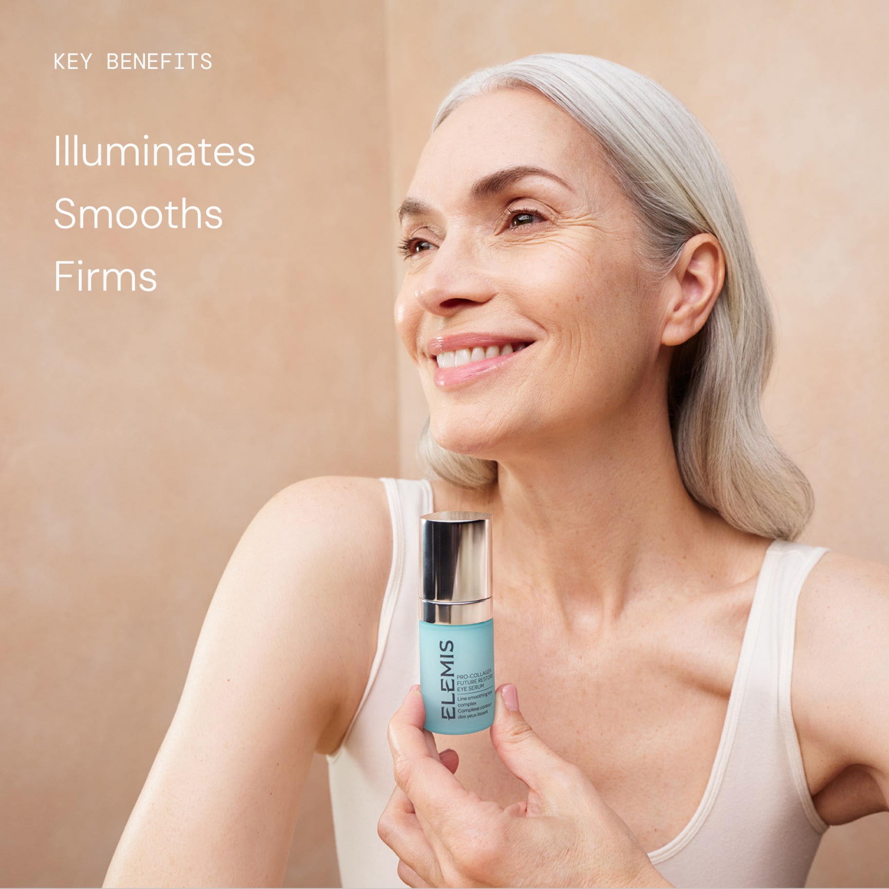 Smiling older woman with long grey hair holds a bottle of ELEMIS skincare serum against a neutral background. Text beside her reads: “Key benefits: Illuminates, Smooths, Firms.”