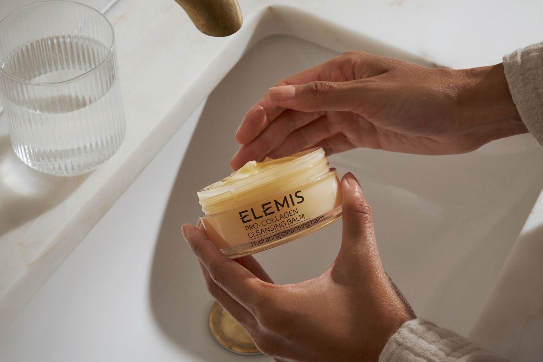 A hand holds three stacked Elemis Pro-Collagen Cleansing Balms (green, yellow, pink) against a bright blue sky and white flowers.