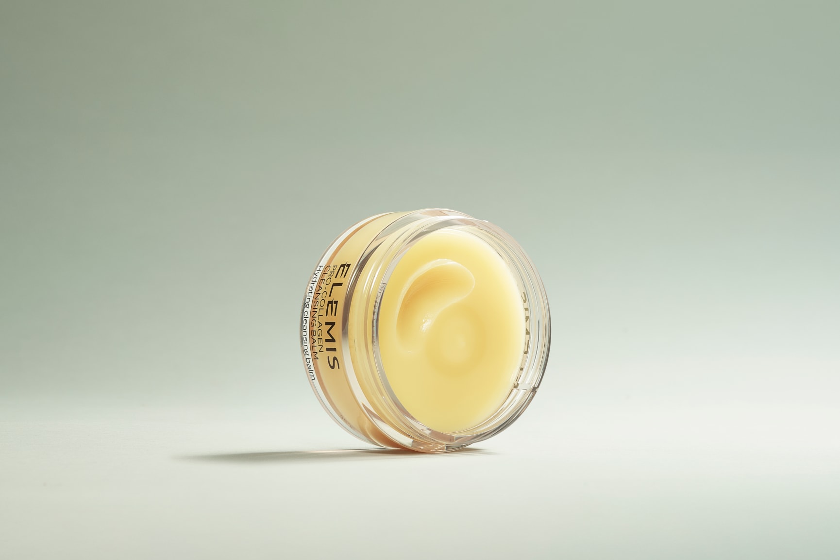 Two Elemis Pro-Collagen skincare products, a mint green cleanser bottle and a yellow cleansing balm jar, sit in shallow water against a dark blue background.