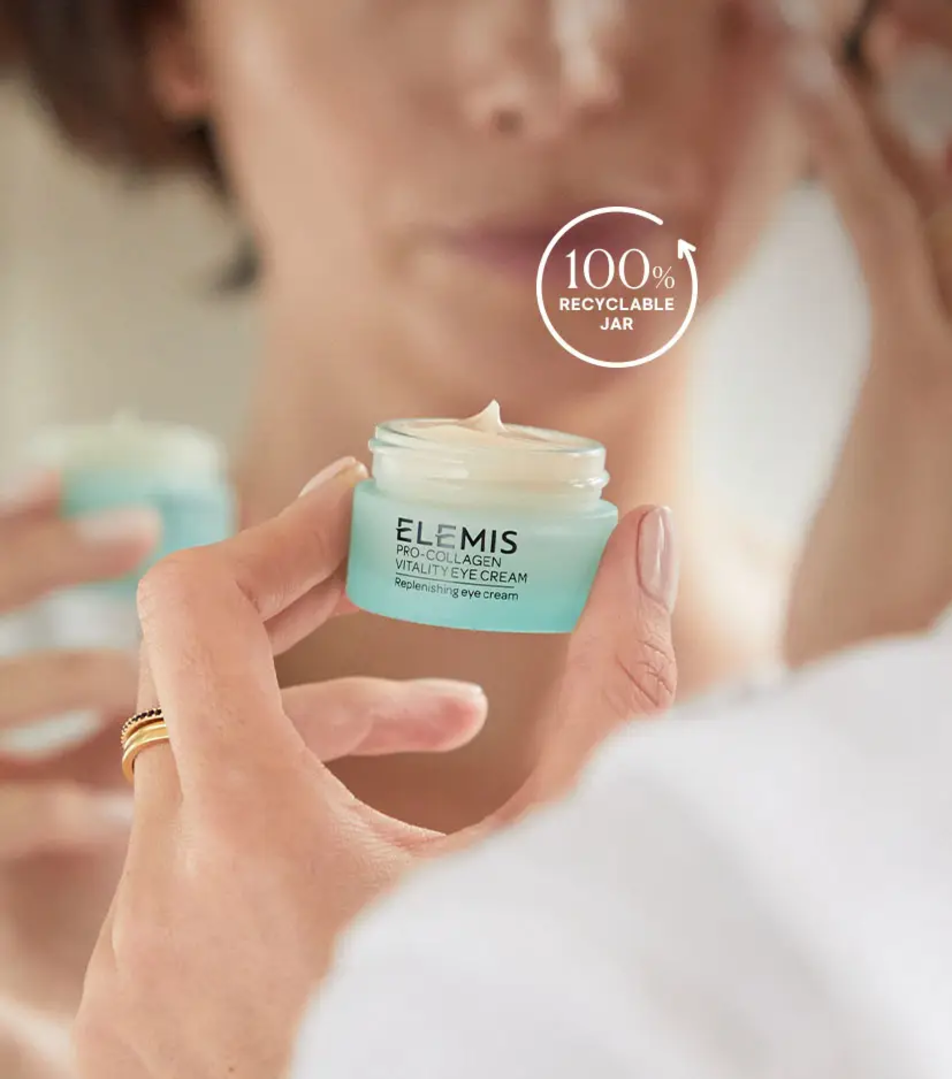 Hand holding open Elemis Pro-Collagen Vitality Eye Cream jar (100% recyclable) with cream visible, reflected in a mirror.