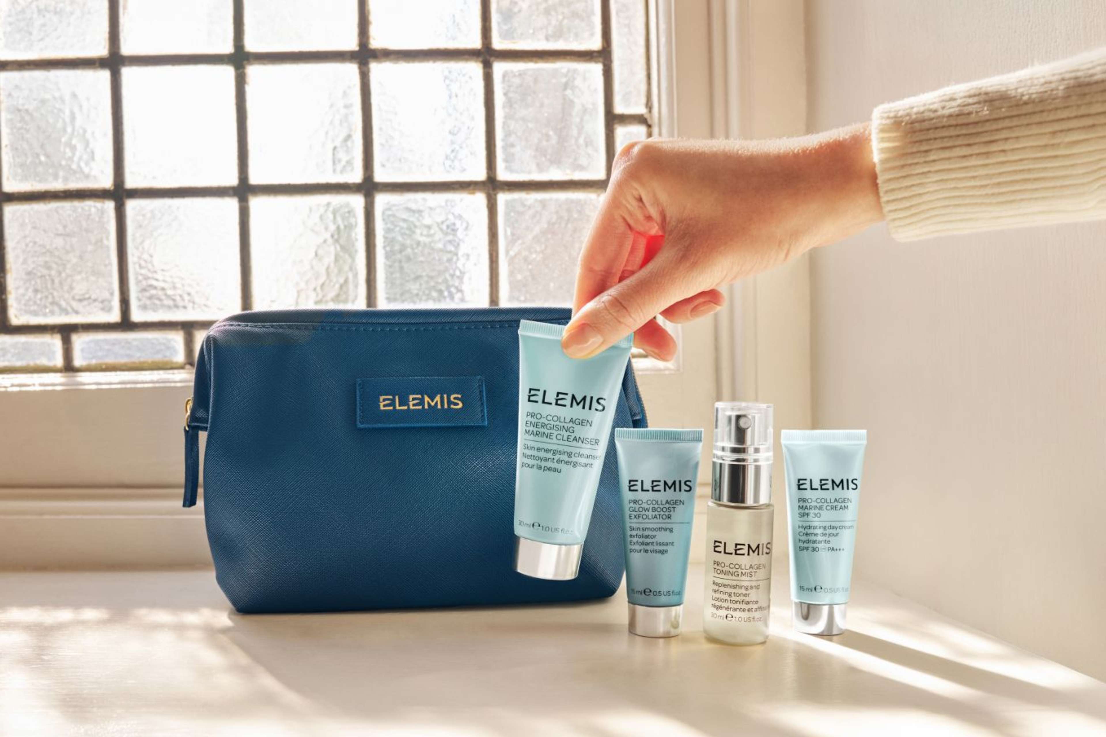 A hand holds an Elemis cleanser next to three other Elemis skincare products and a dark blue Elemis cosmetics bag.