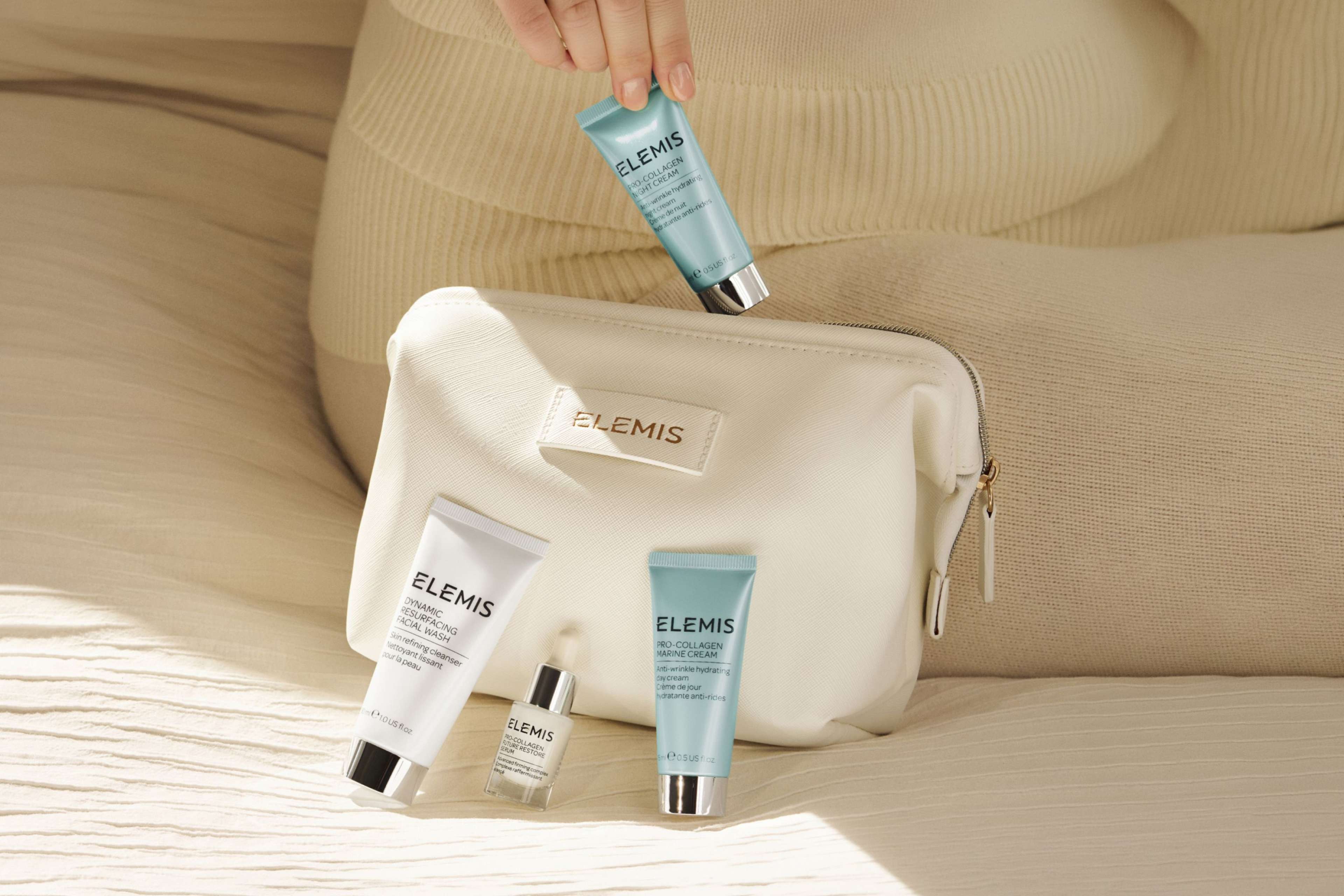 A hand holds an Elemis cleanser next to three other Elemis skincare products and a dark blue Elemis cosmetics bag.