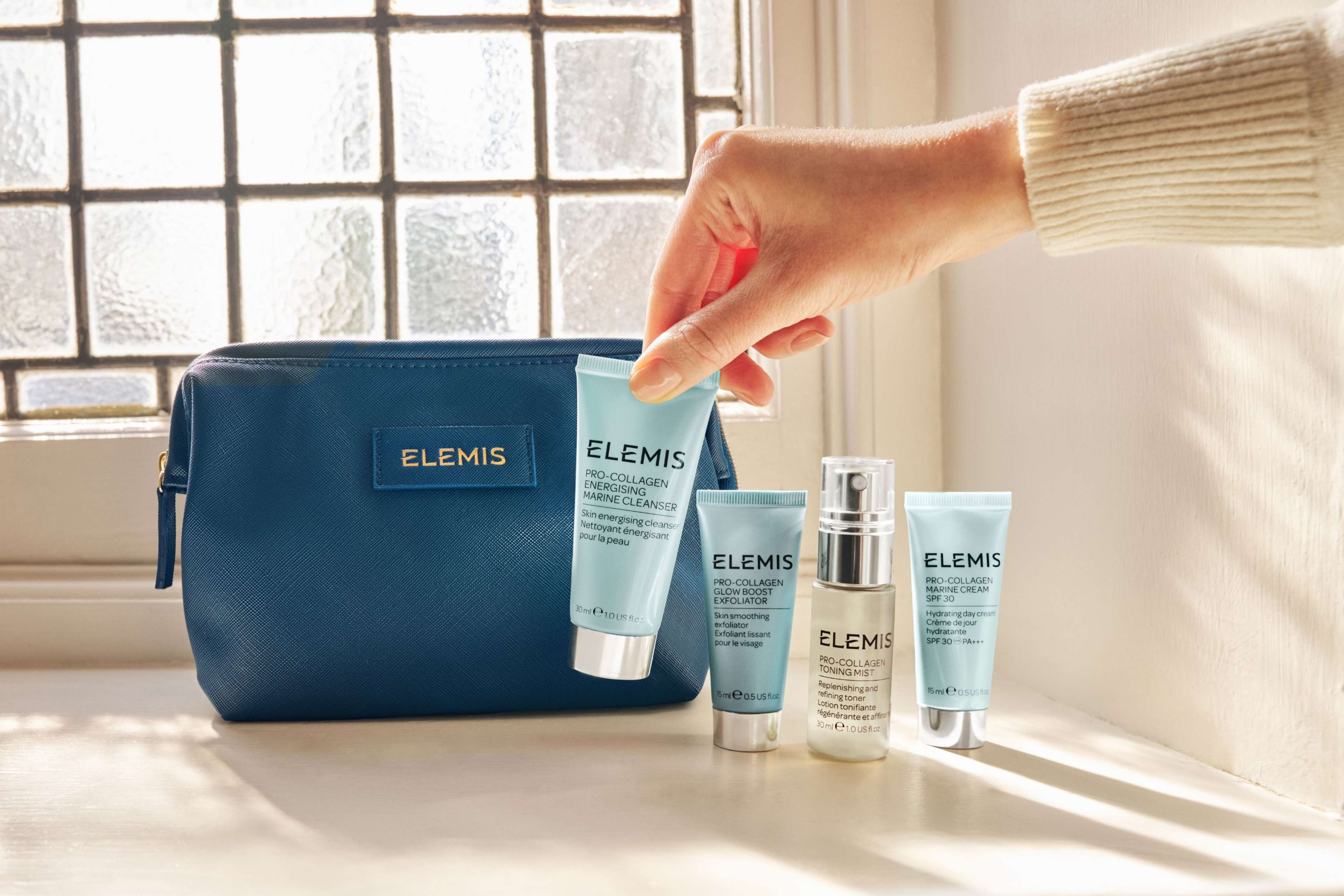 A hand holds an Elemis cleanser next to three other Elemis skincare products and a dark blue Elemis cosmetics bag.