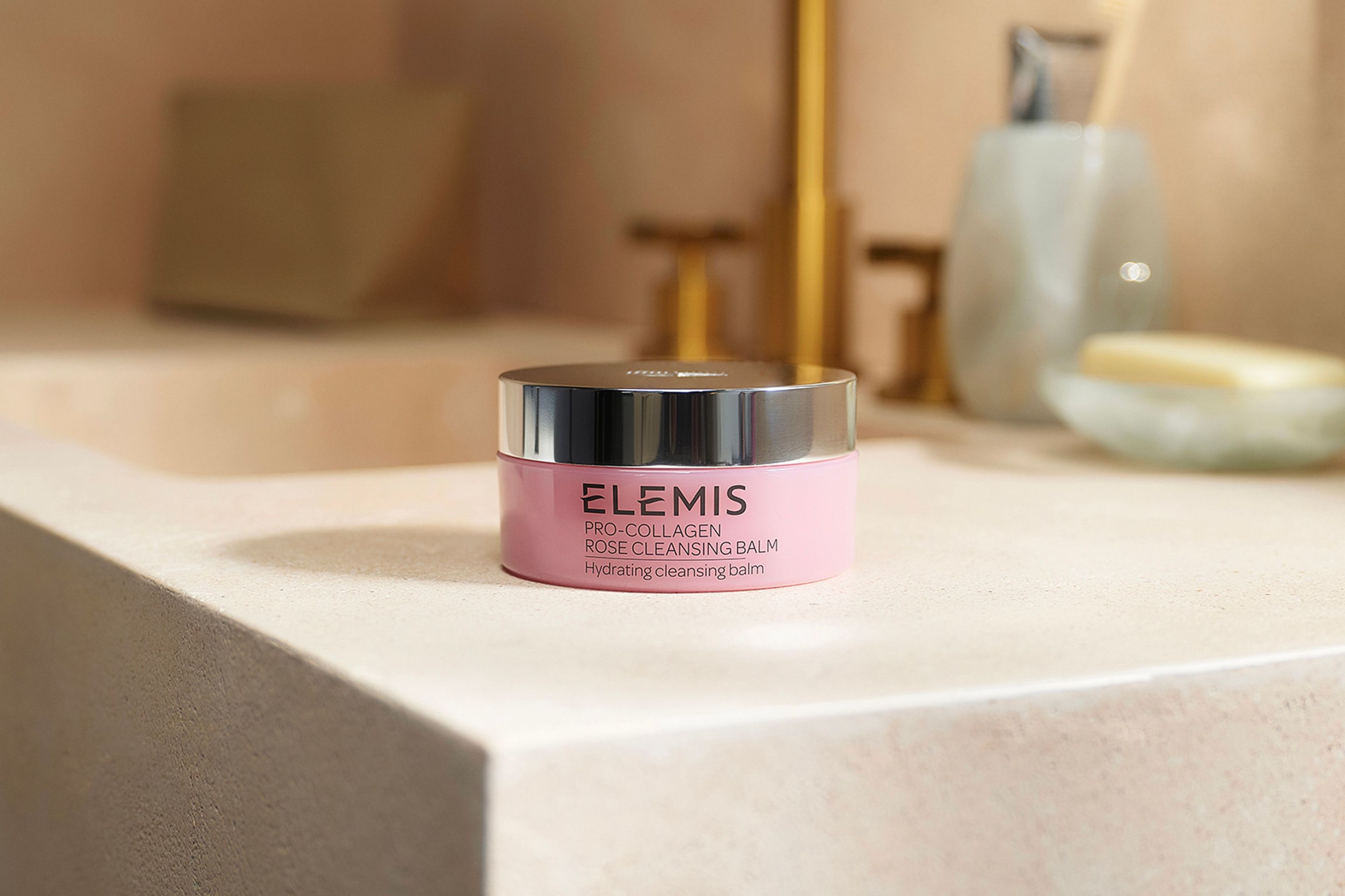 A pink jar of Elemis Pro-Collagen Rose Cleansing Balm sits in rippling water next to a pink rose.