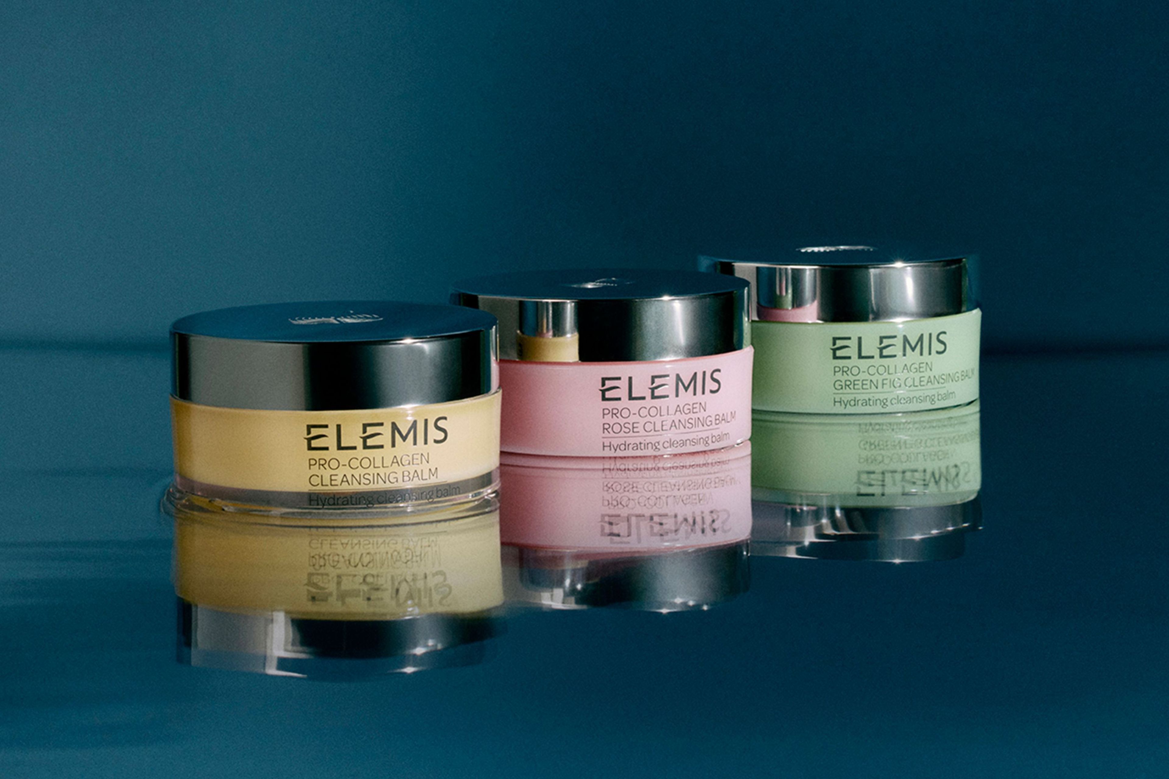 Una mano tiene tre vasetti di ELEMIS Pro-Collagen Cleansing Balm impilati (verde, oro, rosa) su uno sfondo di cielo azzurro con fiori di campo.