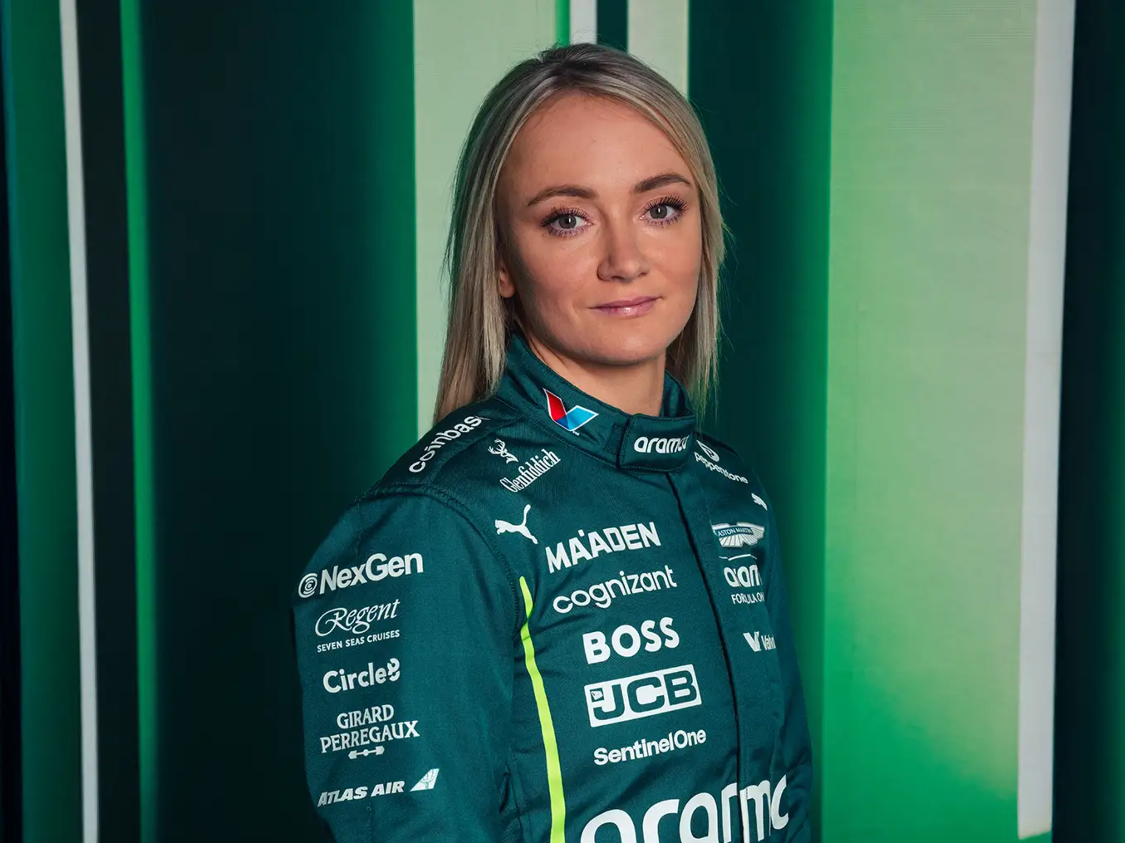 Una mujer rubia con un traje de carreras verde oscuro adornado con logotipos de patrocinadores sonríe levemente, mirando directamente al espectador, sobre un fondo verde a rayas.