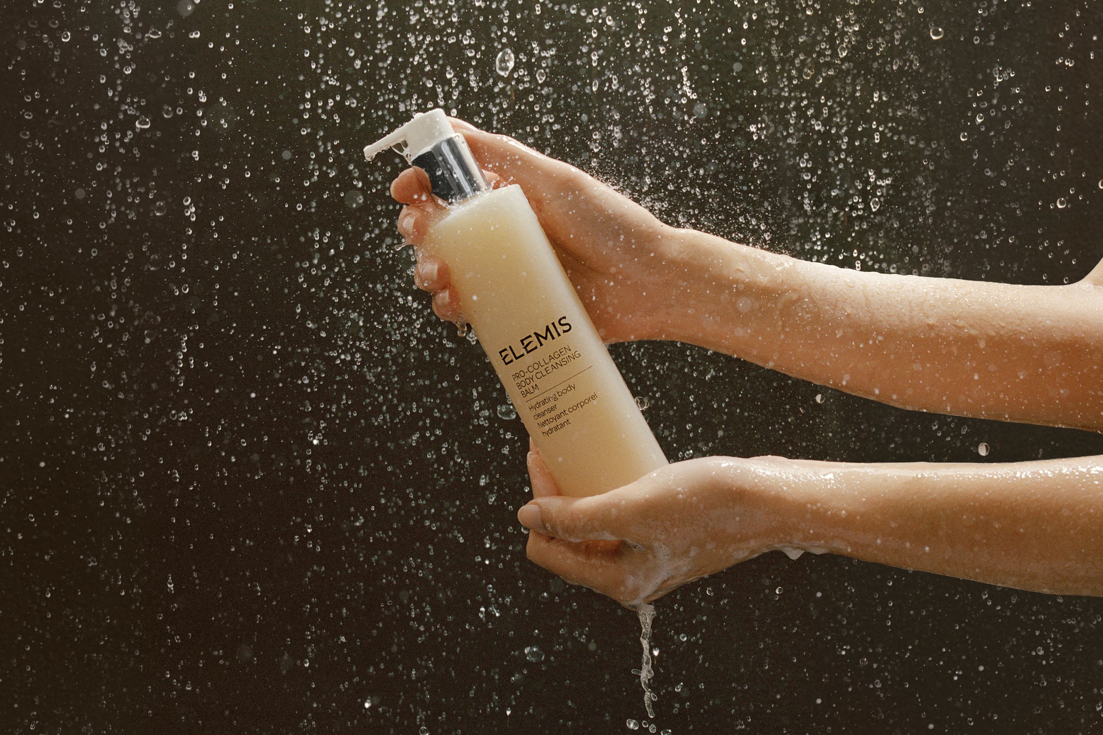 Des mains tenant une bouteille de Pro-Collagen Body Cleansing Balm d’ELEMIS sous un jet d’eau.