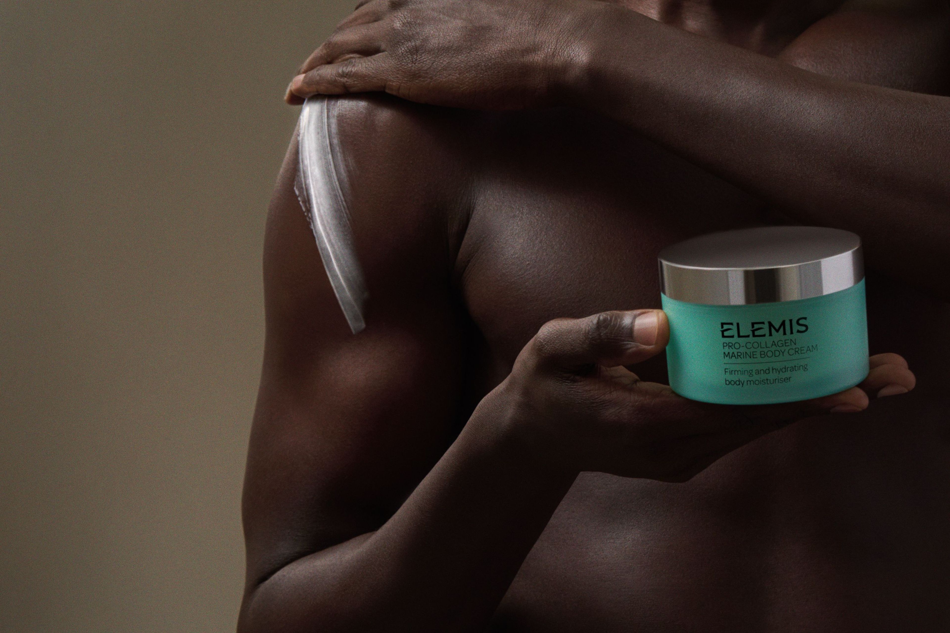 A person with dark skin holds a teal Elemis body cream jar, with a white streak of cream on their shoulder.