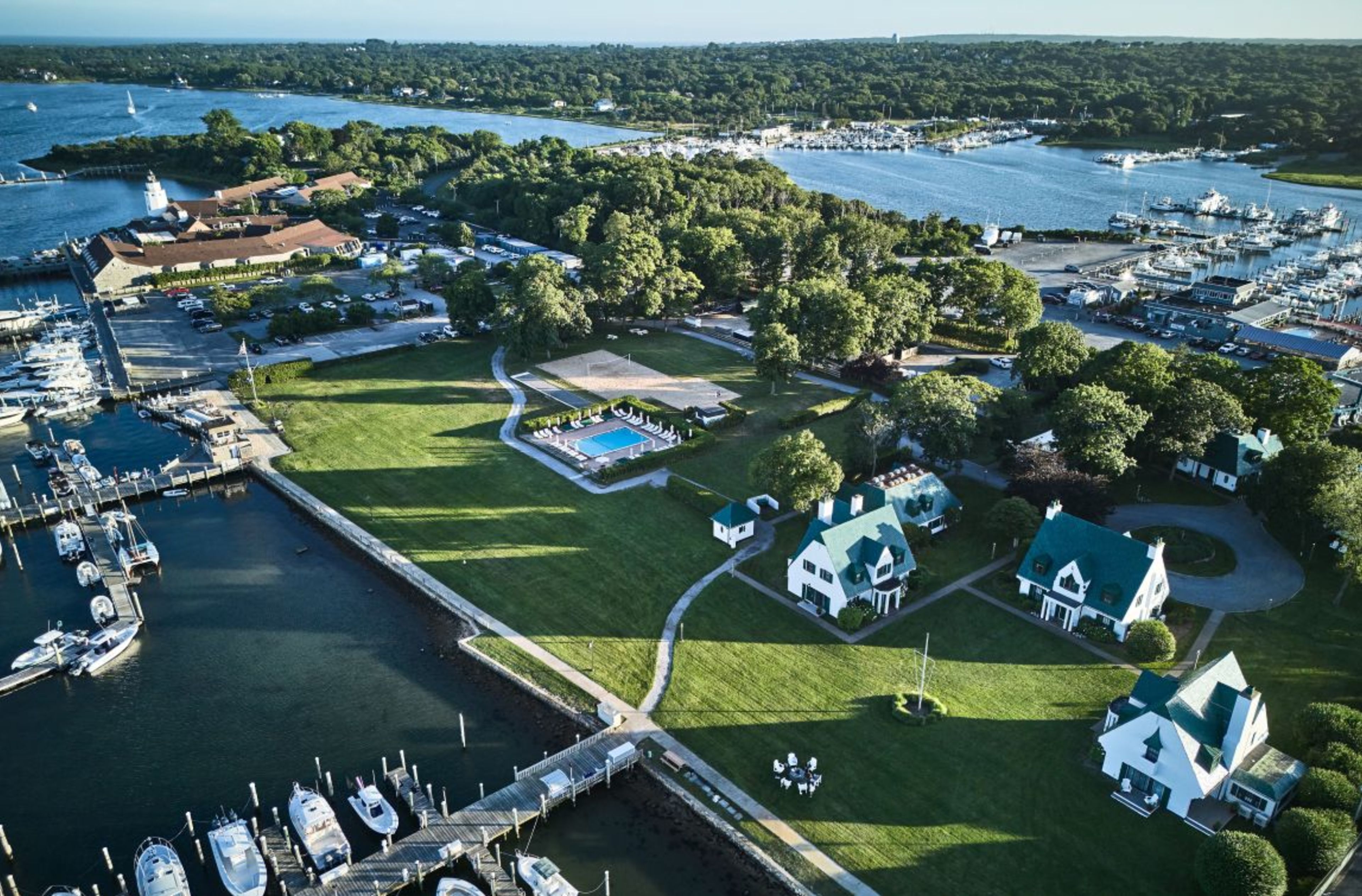 Aerial view of a sprawling waterfront resort with white buildings, green roofs, a pool, volleyball court, extensive lawns, and multiple boat-filled marinas.