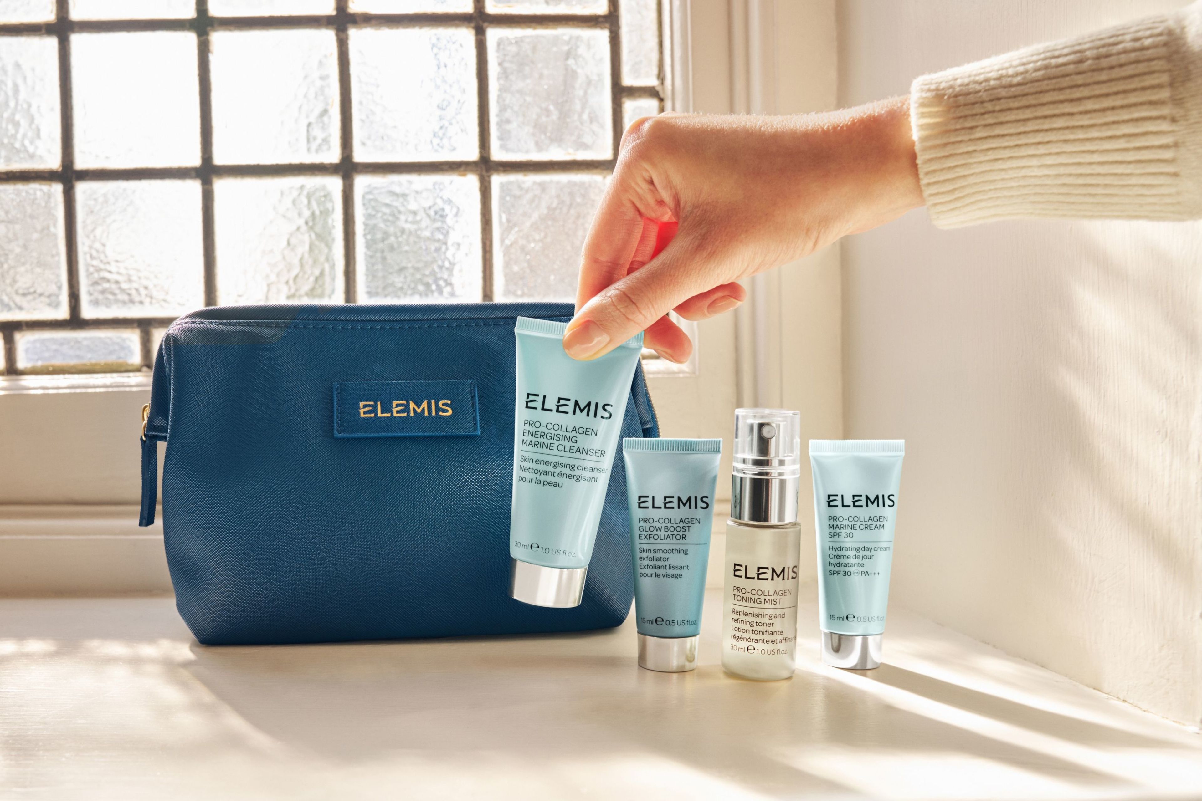 A hand holds an Elemis cleanser next to three other Elemis skincare products and a dark blue Elemis cosmetics bag.