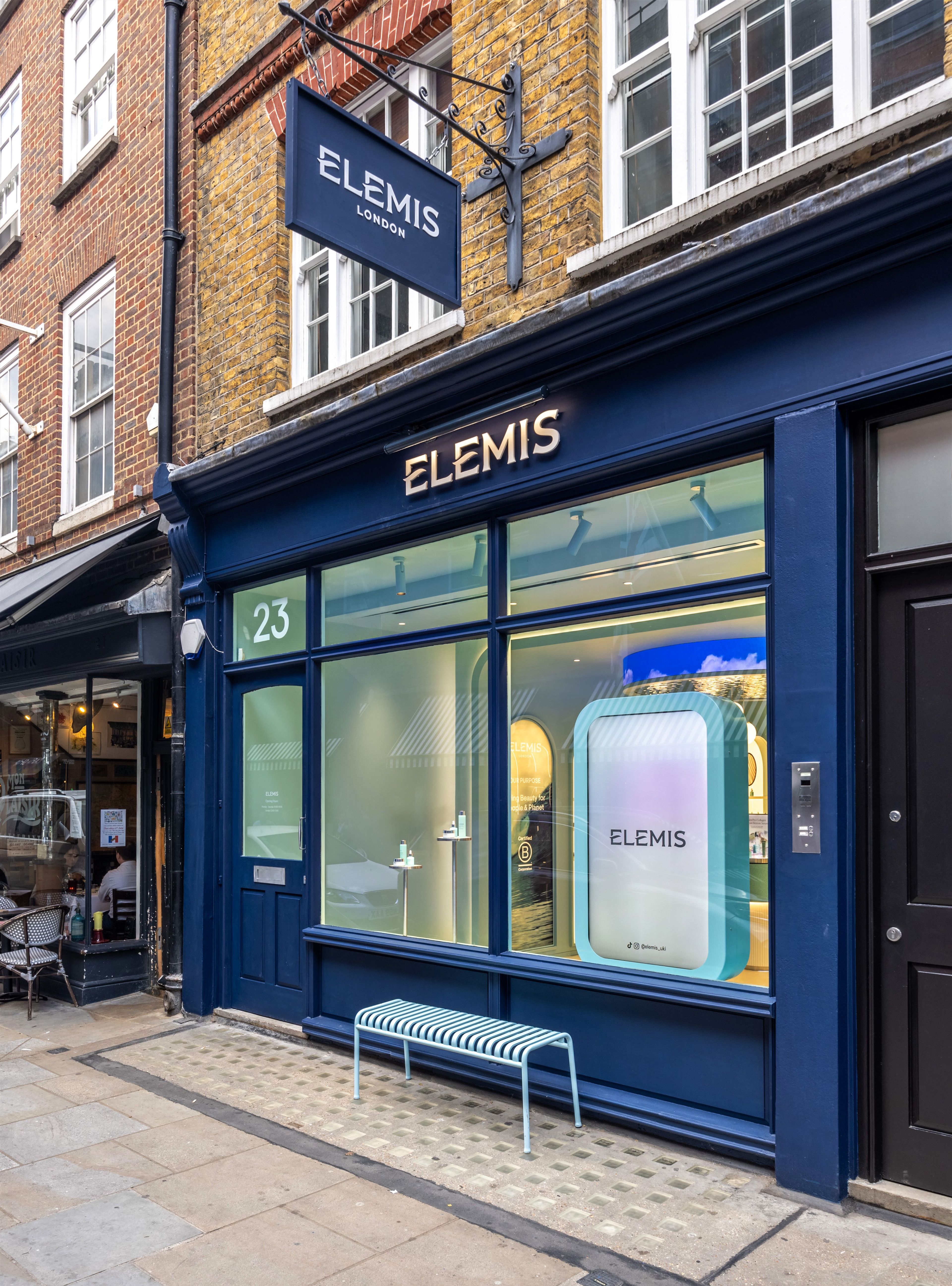 A dark blue Elemis storefront with a hanging sign, illuminated name above the windows, and a light blue bench on the sidewalk.