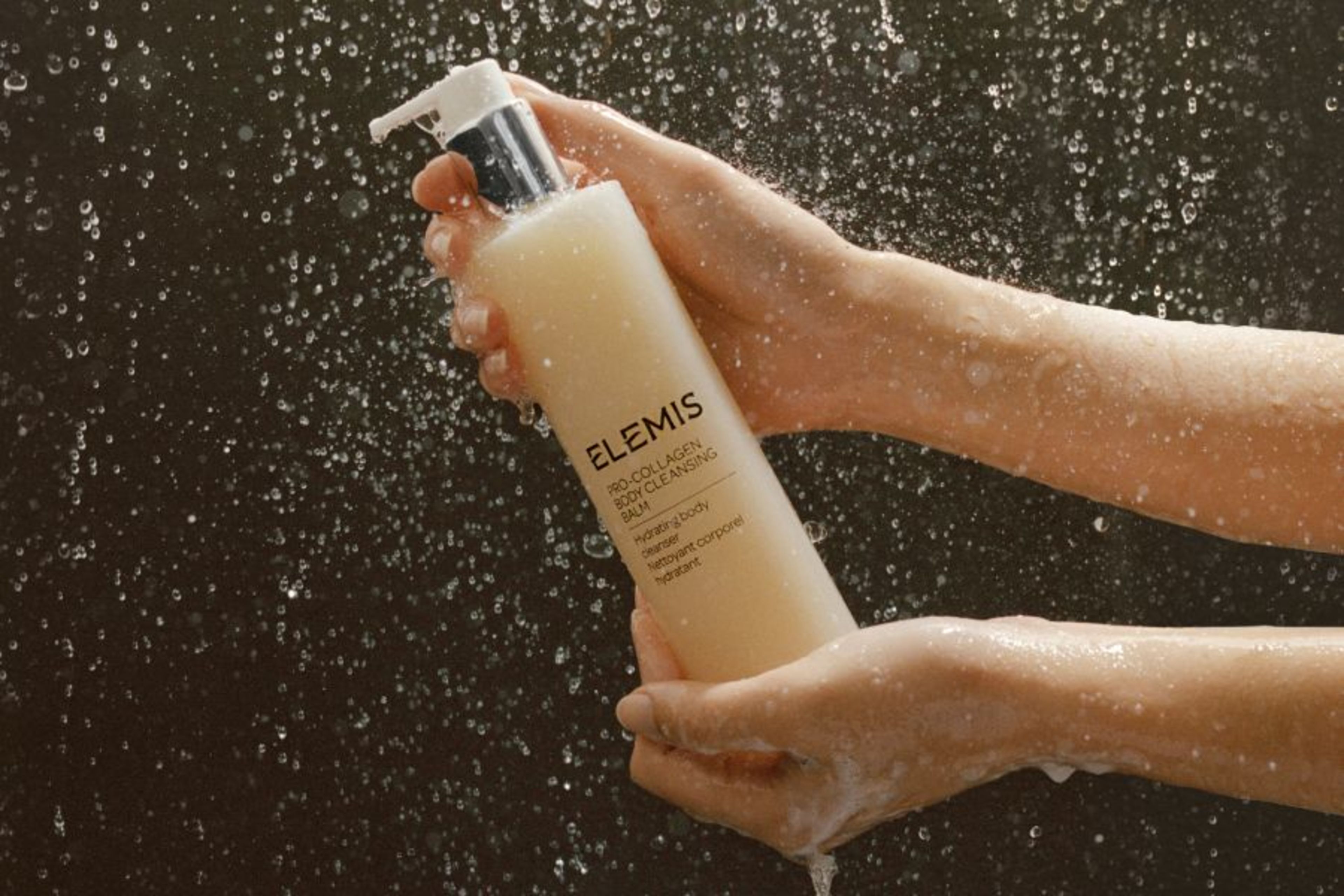 Hands holding an Elemis Pro-Collagen Body Cleansing Balm bottle under falling water droplets.