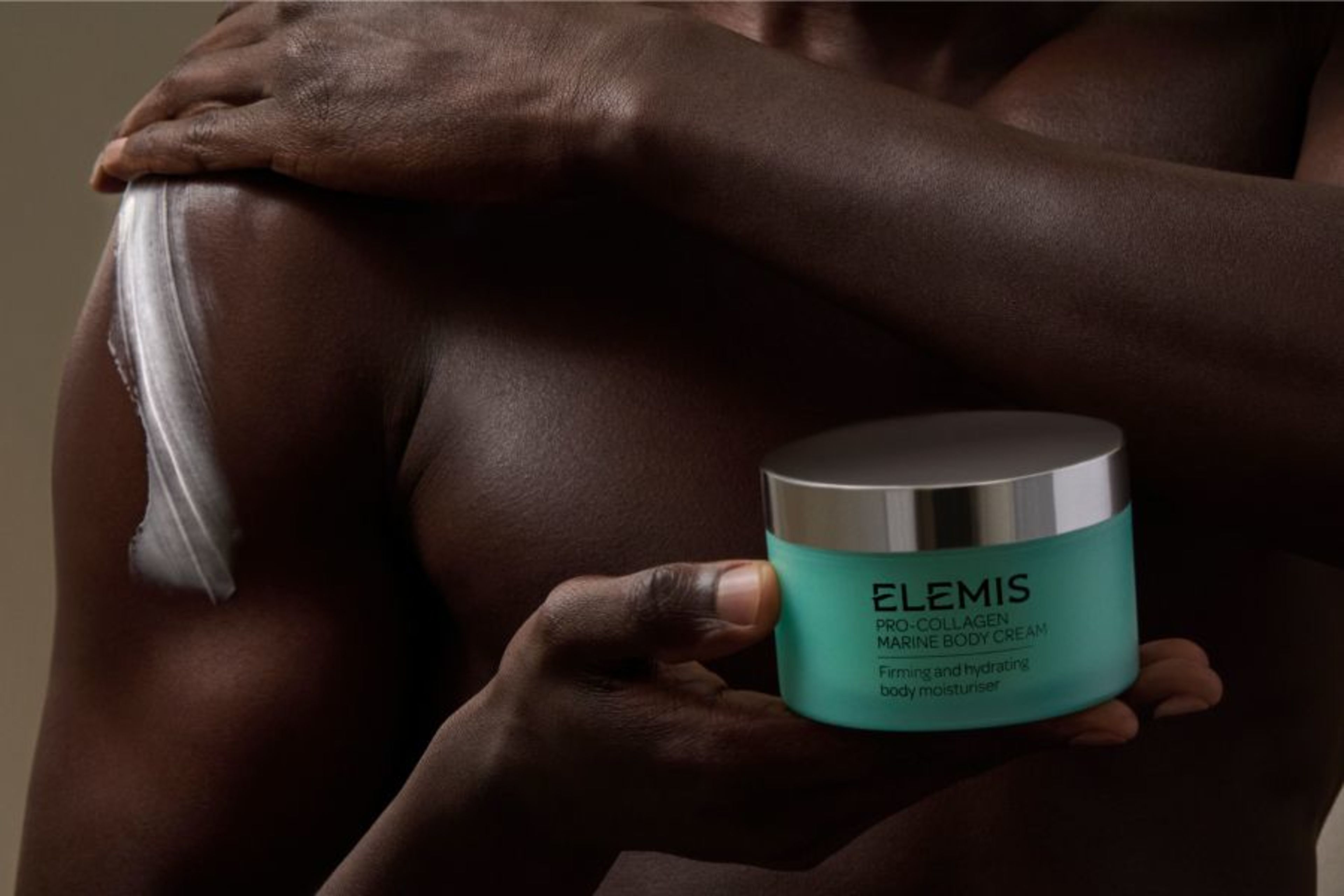 A person with dark skin applies Elemis Pro-Collagen Marine Body Cream to their shoulder.