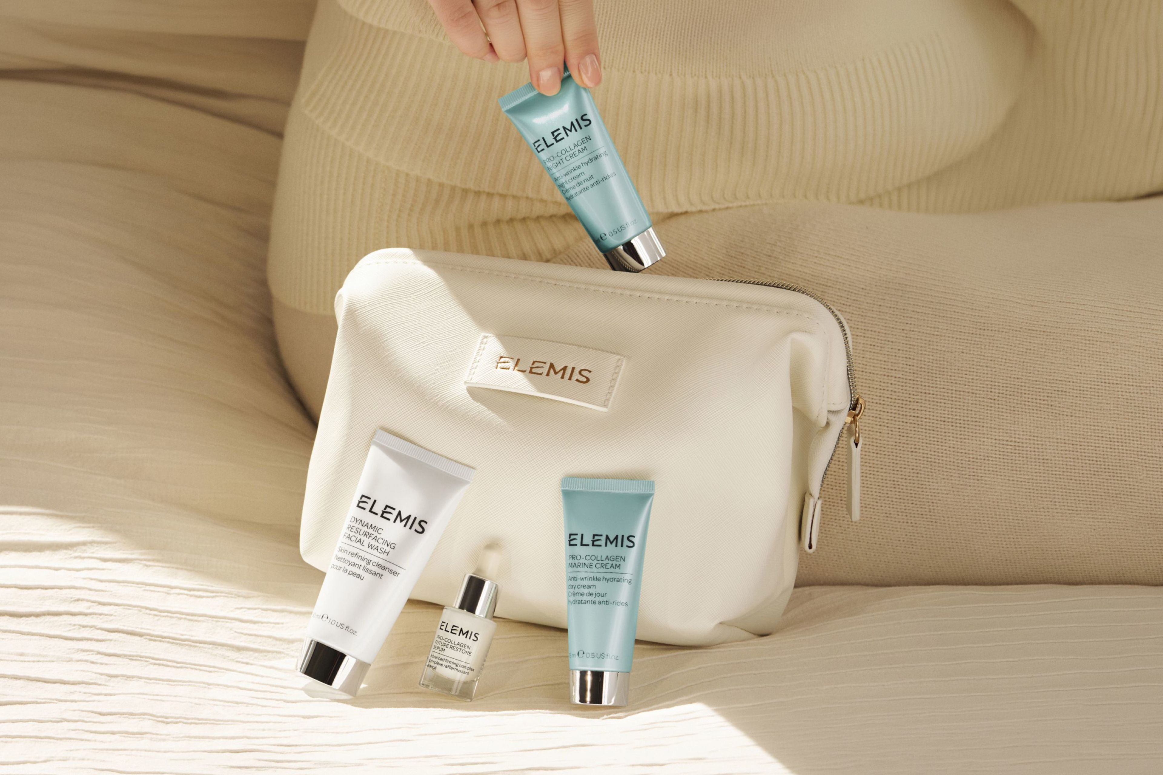 A hand holds an Elemis cleanser next to three other Elemis skincare products and a dark blue Elemis cosmetics bag.