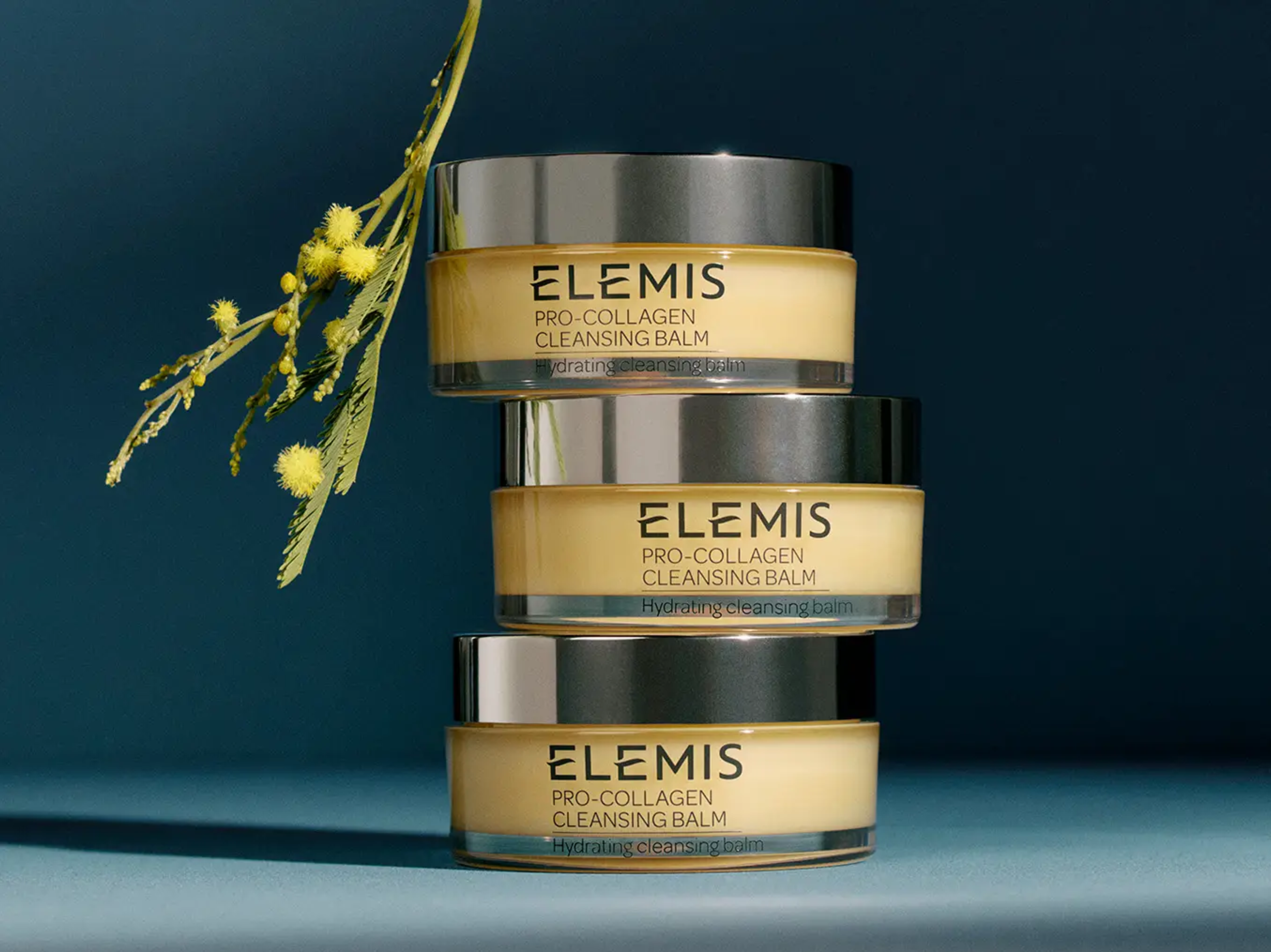 Tre barattoli impilati di Elemis Pro-Collagen Cleansing Balm accanto a un rametto di fiori di mimosa gialla.