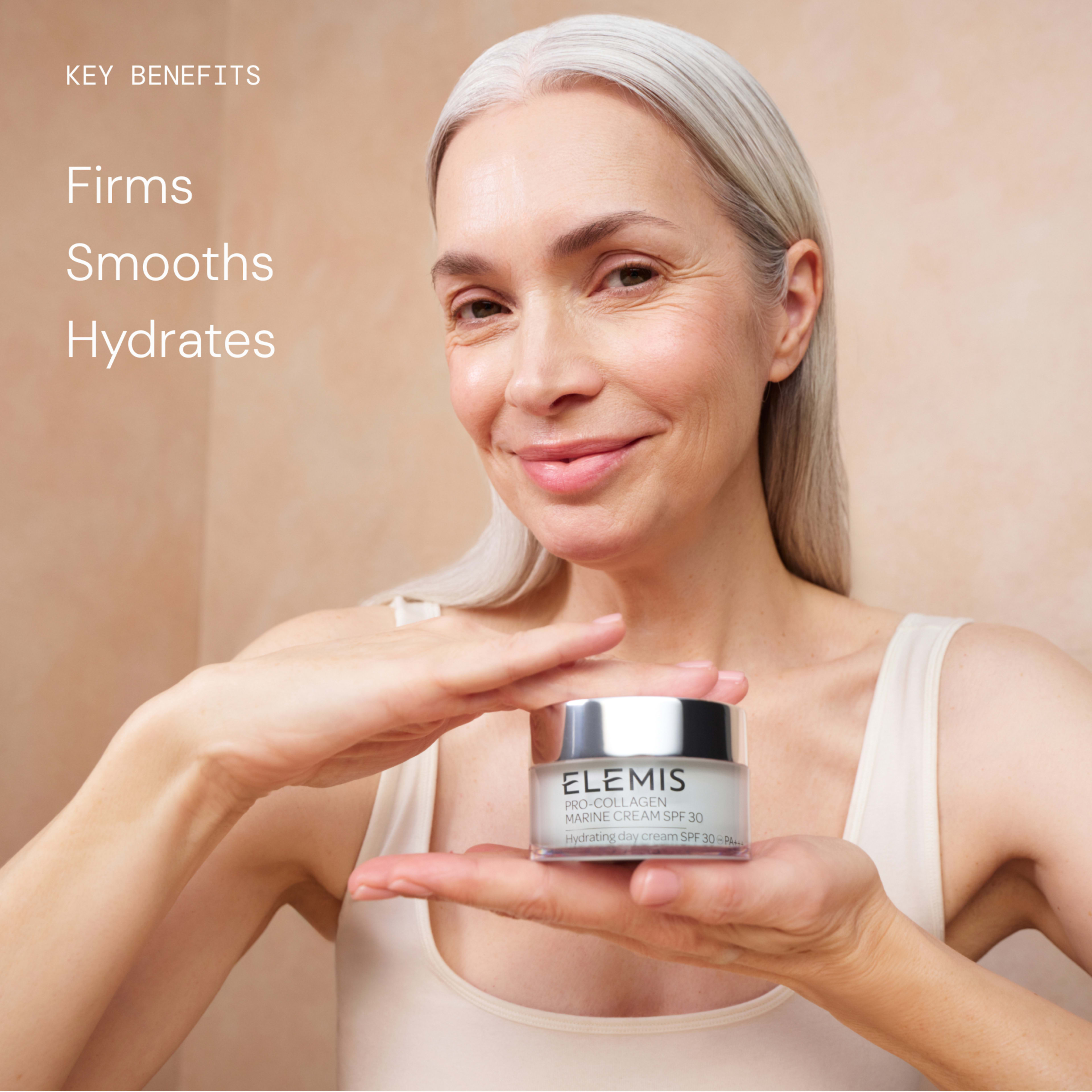 Smiling woman with long grey hair presents a jar of ELEMIS Pro-Collagen Marine Cream SPF30, framed between her hands against a neutral background. On the left, text reads: “Key benefits – Firms, Smooths, Hydrates.”
