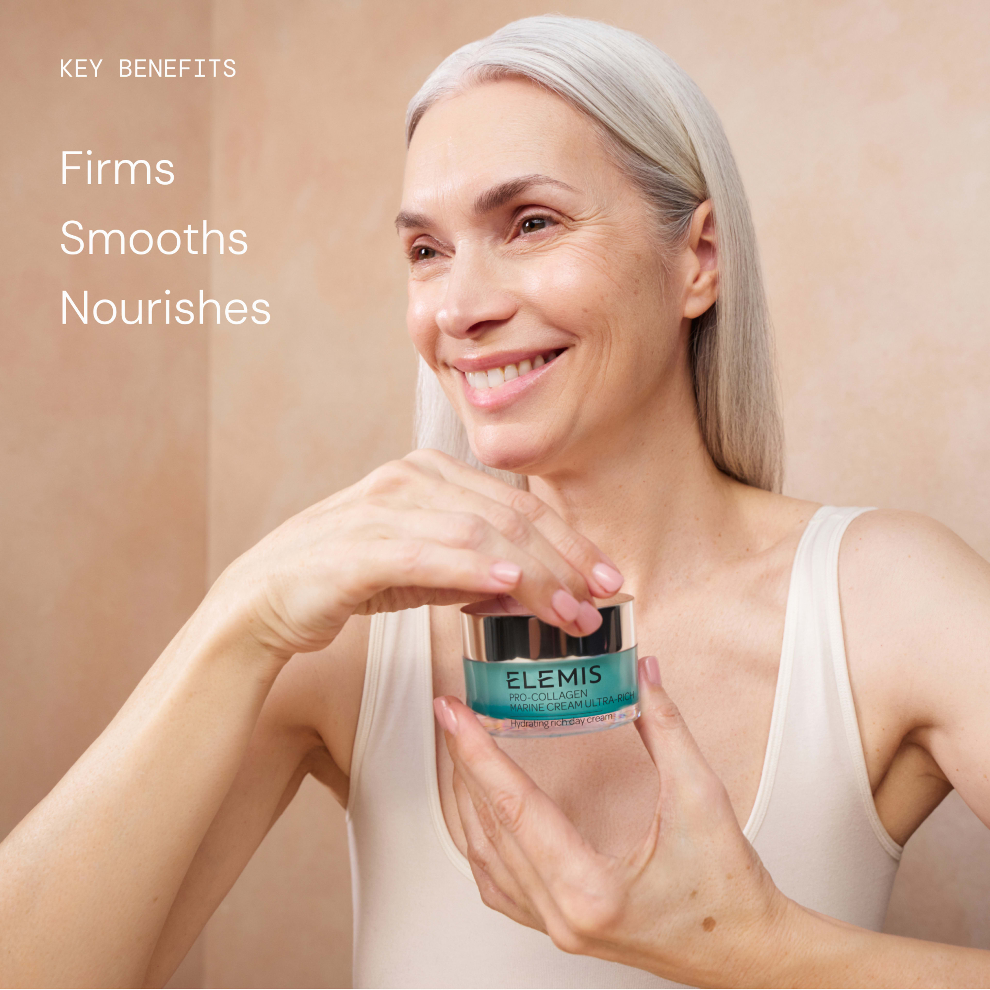Smiling woman with long grey hair applies ELEMIS Pro-Collagen Marine Cream from a jar, wearing a sleeveless top against a neutral bathroom-style background. On the left, text reads: "Key benefits: Firms, Smooths, Nourishes".