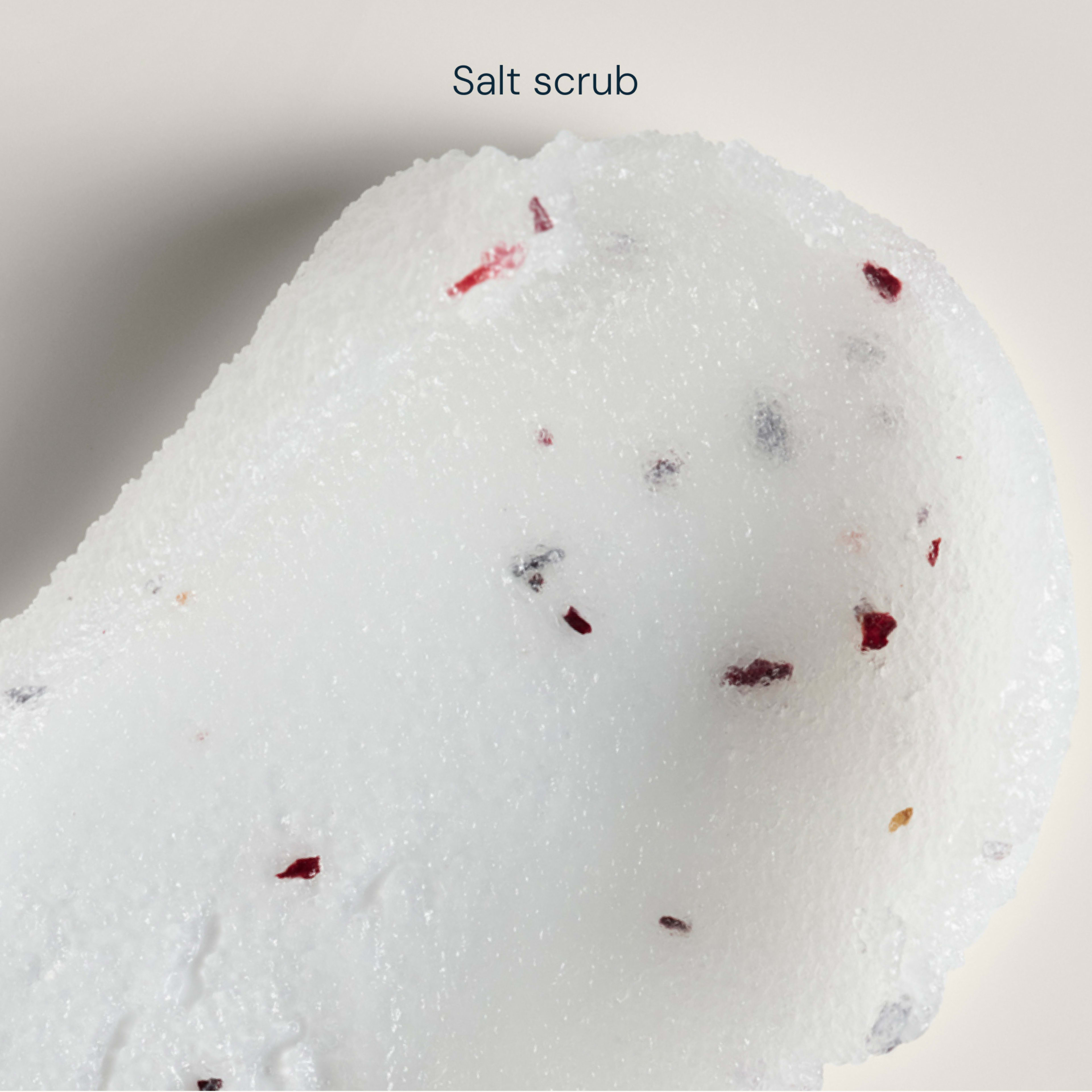 Close-up swatch of white salt scrub with a soft, granulated texture and small red and dark exfoliating particles against a pale background, with the words "Salt scrub" above.