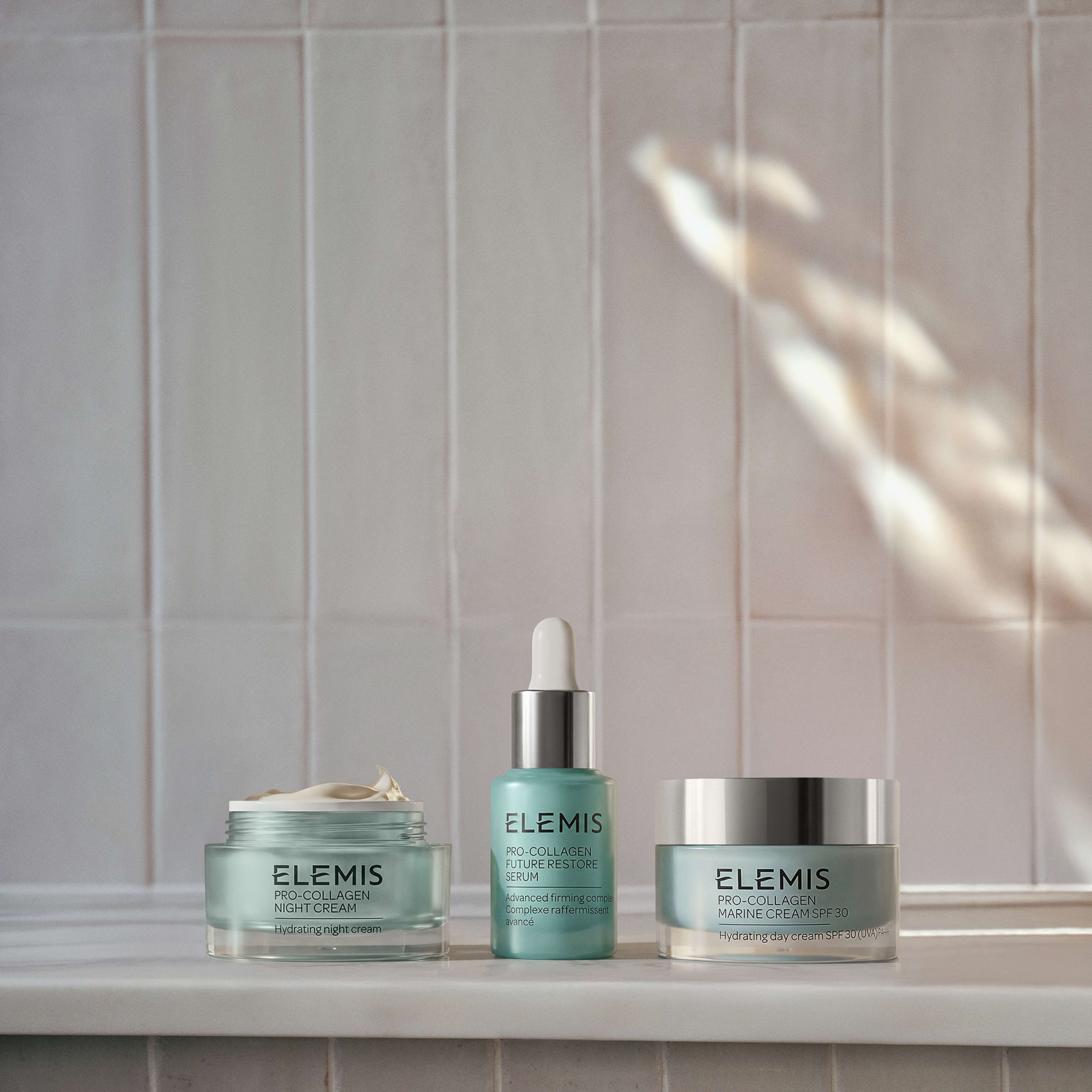 Three ELEMIS Pro-Collagen products arranged on a bathroom ledge against pale tiled wall: an open Pro-Collagen Night Cream jar, Pro-Collagen Elixir serum with dropper, and closed Pro-Collagen Marine Cream SPF jar, lit by soft natural light.