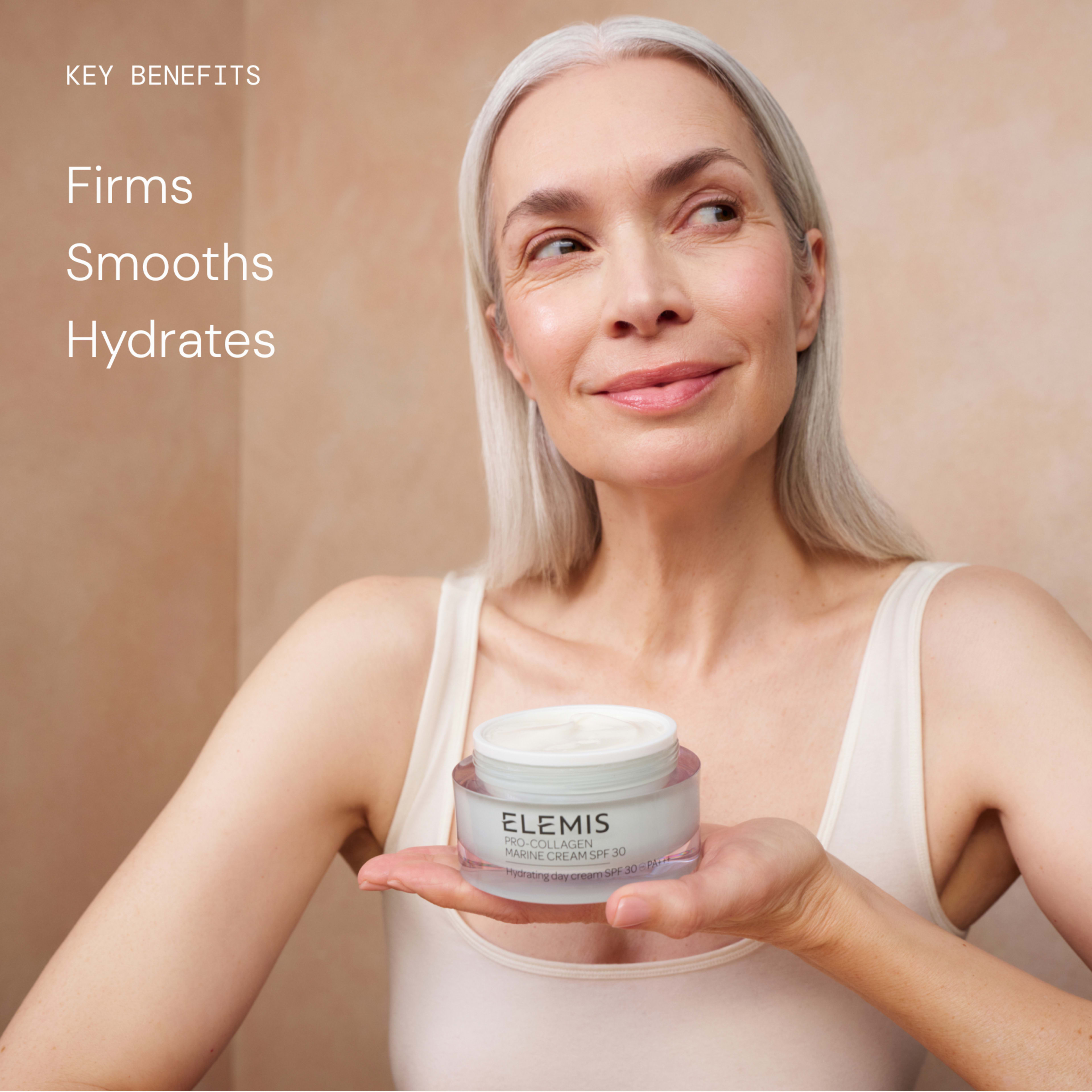 Smiling woman with minimal makeup applies eye cream while holding an open jar of ELEMIS Pro-Collagen Marine Cream SPF30, showing its texture. Text reads: "Key benefits: Firms, Smooths, Hydrates".