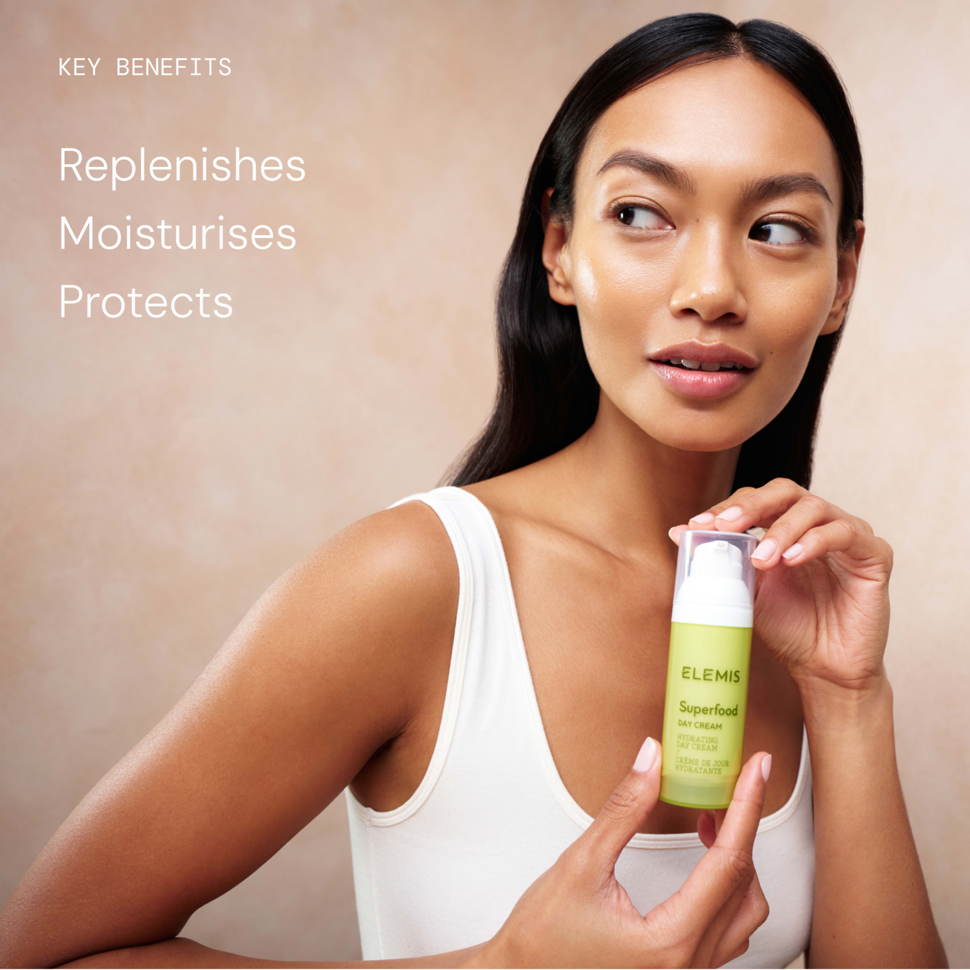 Woman in a white vest holding an ELEMIS Superfood Day Cream bottle, looking to the side against a neutral background. On the left, text reads: "Key benefits: Replenishes, Moisturises, Protects".