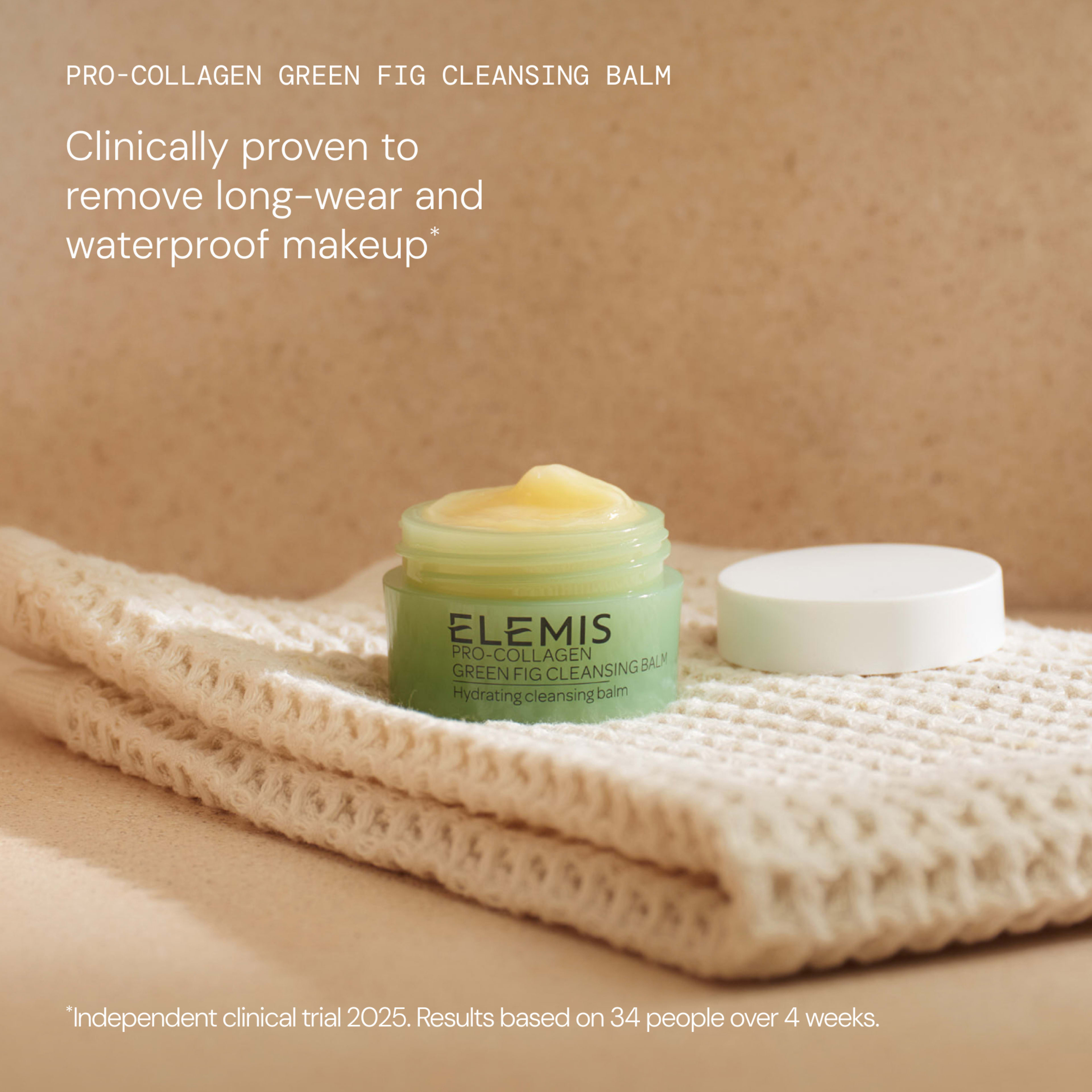 ELEMIS Pro-Collagen Green Fig Cleansing Balm jar open on folded cream towel with lid beside it, against a beige background. On-image text states it is “clinically proven to remove long-wear and waterproof makeup” with an independent clinical trial note.