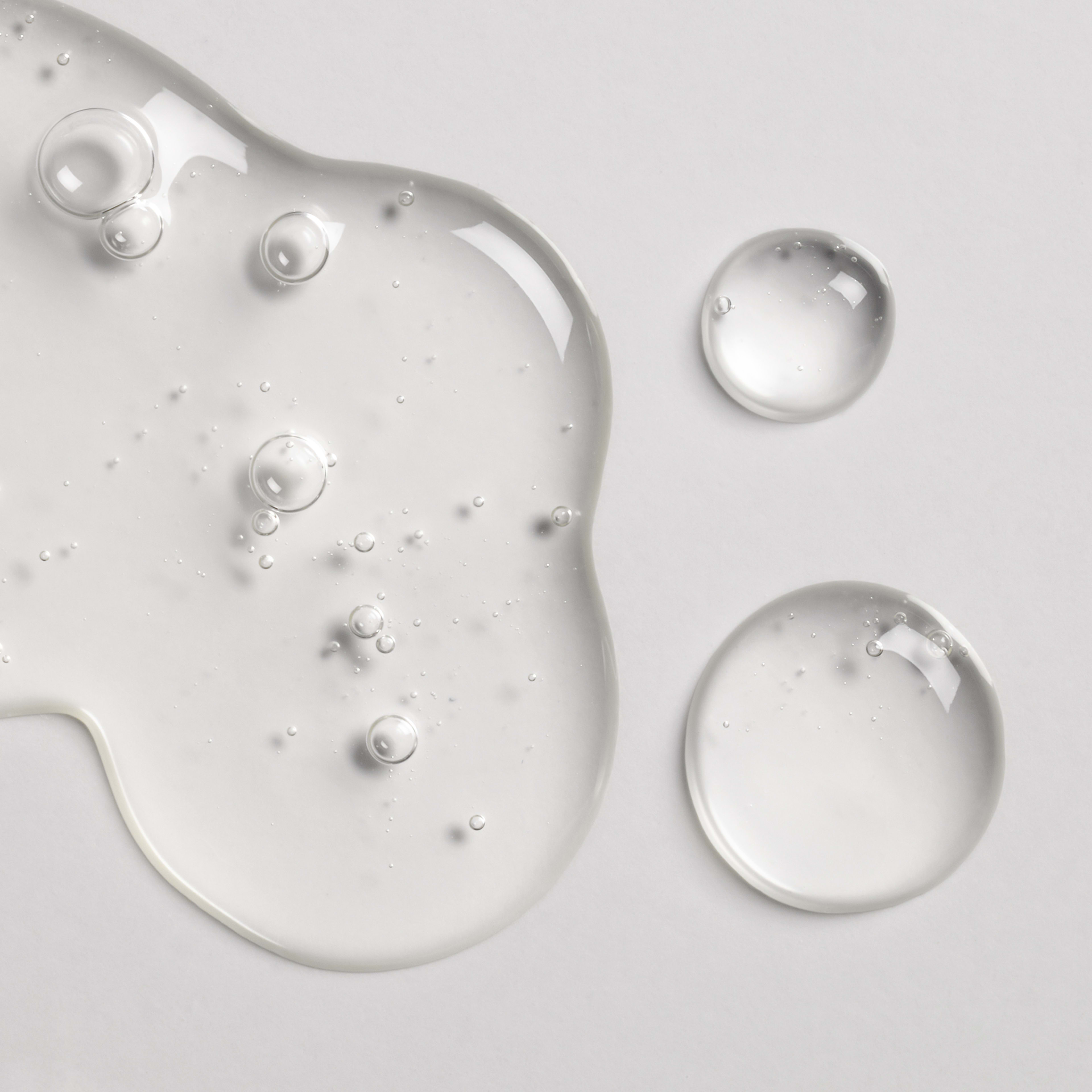 Clear gel skincare serum swatches on a light grey background, showing glossy texture and tiny air bubbles.