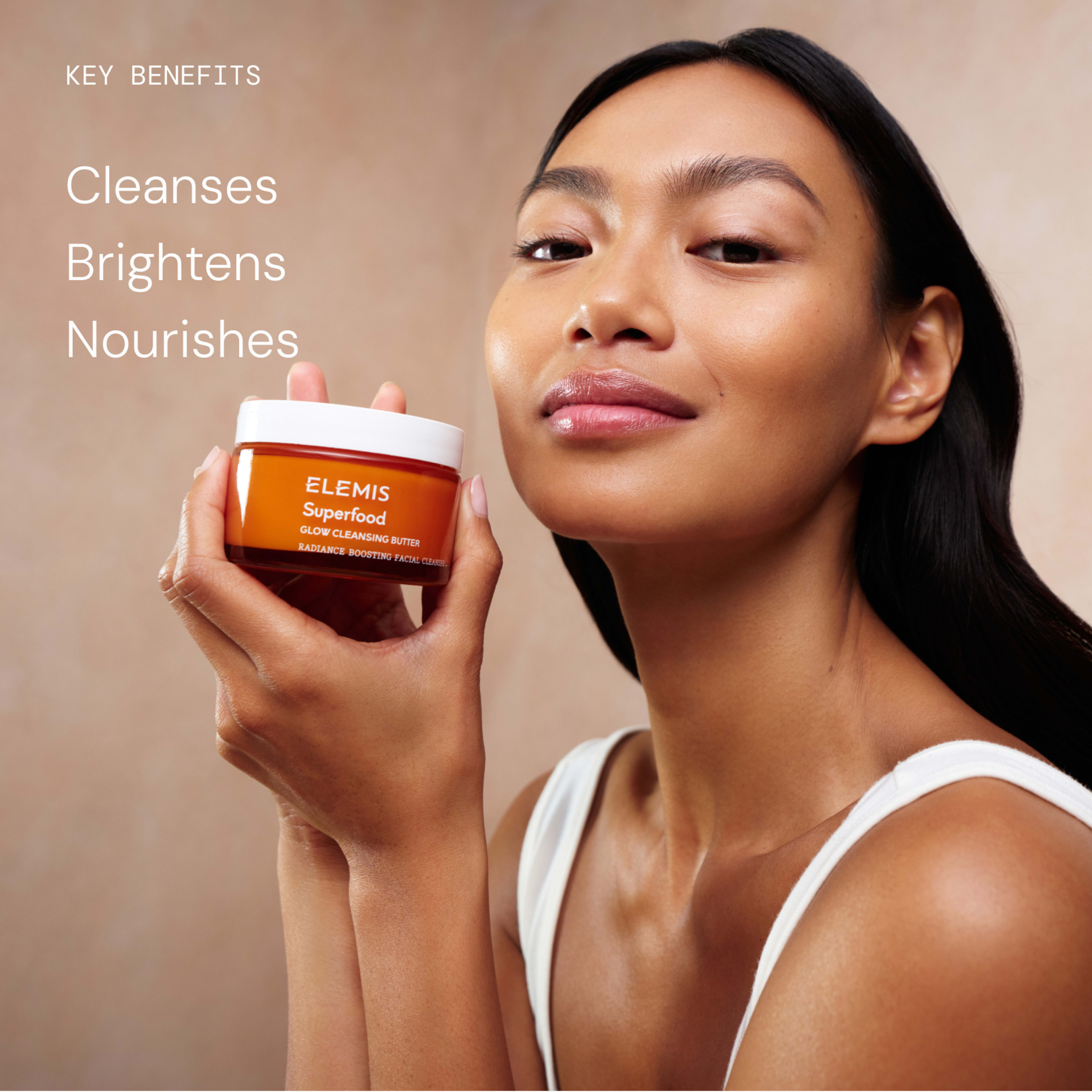 Woman with glowing skin smiles while holding an ELEMIS Superfood Glow Cleansing Butter jar beside her face. Text on the left reads: “Key benefits: Cleanses, Brightens, Nourishes.”